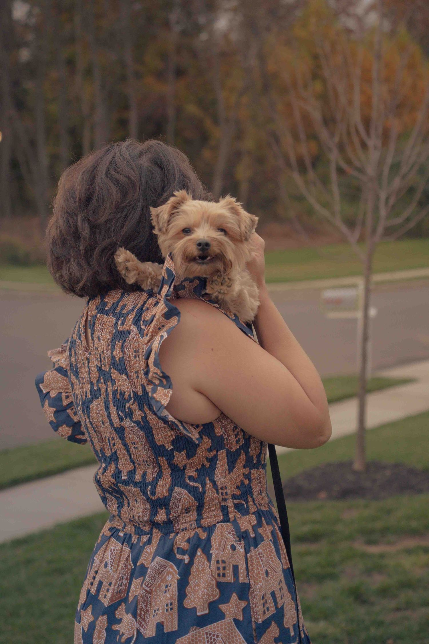 A woman in a blue floral dress cuddles a small Yorkshire Terrier over her shoulder outdoors.