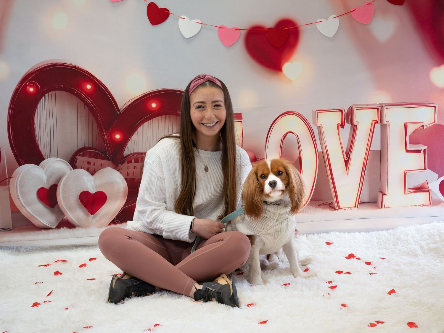 Young woman sits cross-legged with Cavalier King Charles Spaniel in Valentine's Day themed photo with LOVE sign.