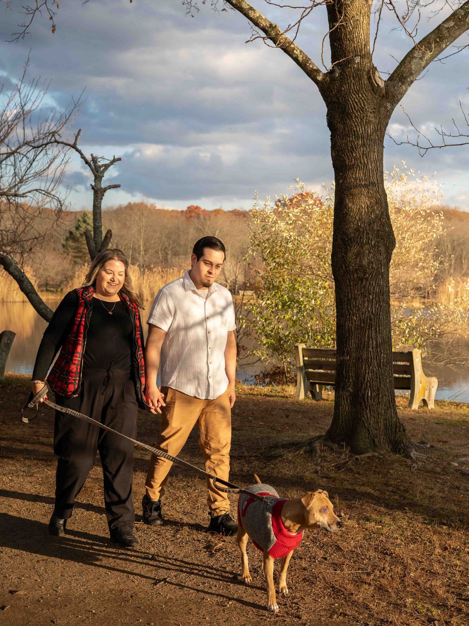 Couple walking their dog on a leash through a sunlit autumn park path near a lake.