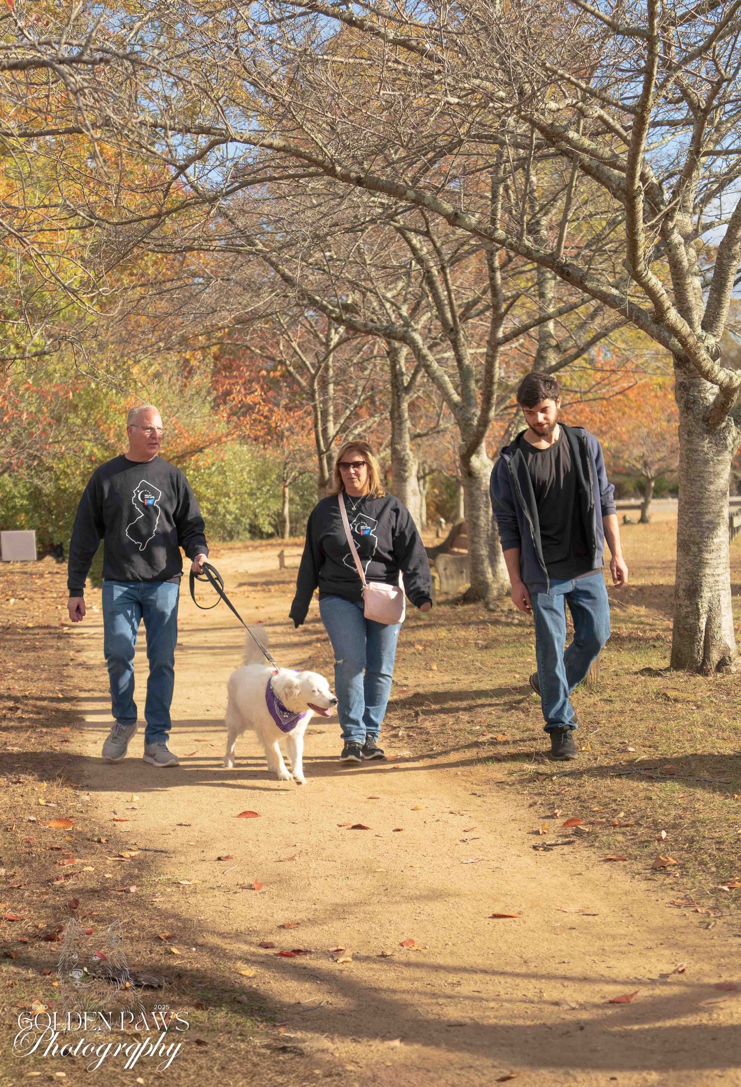 Three people walking a white dog on a leash along a park path surrounded by autumn trees.