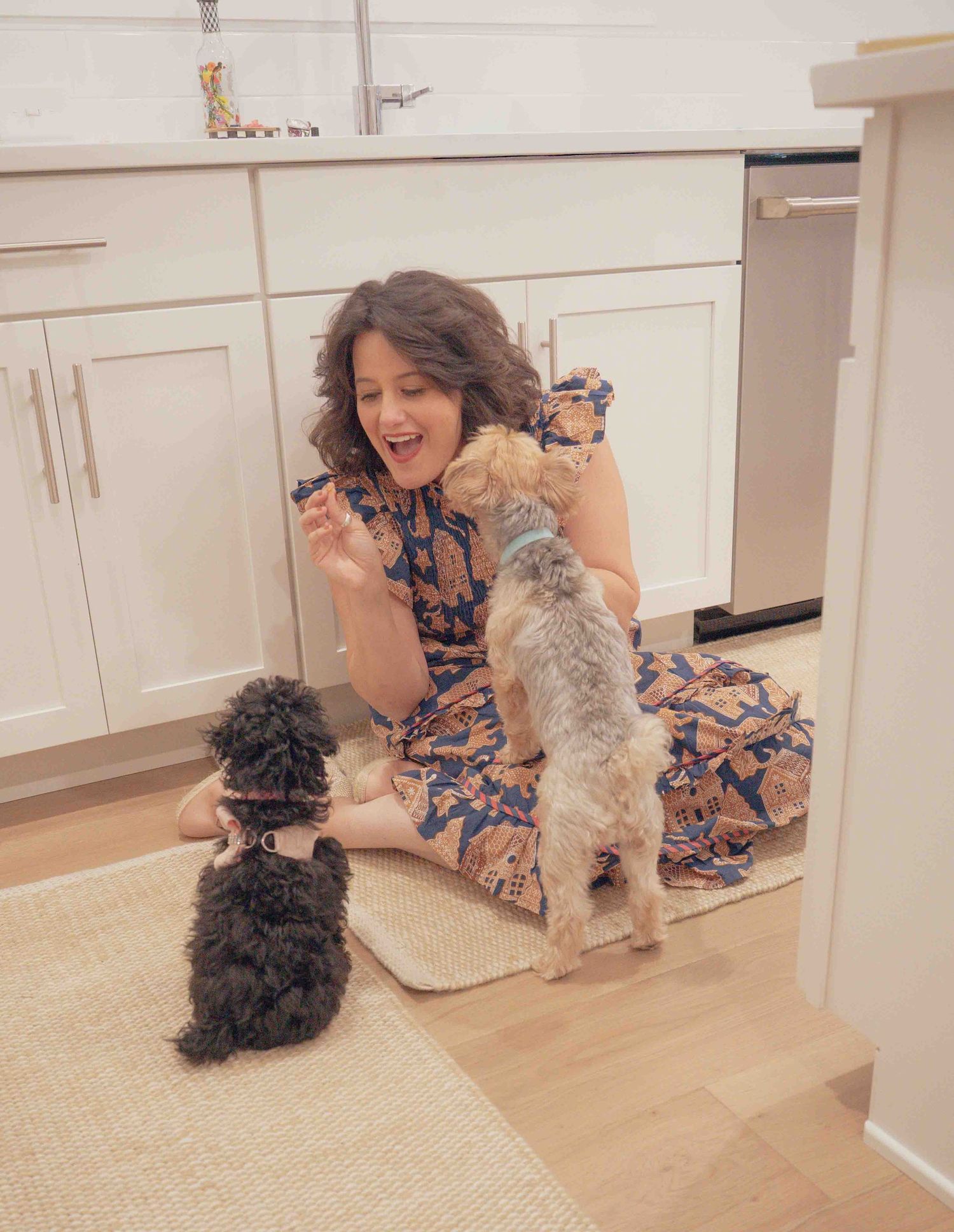 A woman in a floral dress sits on a kitchen floor laughing while three small dogs play around her.