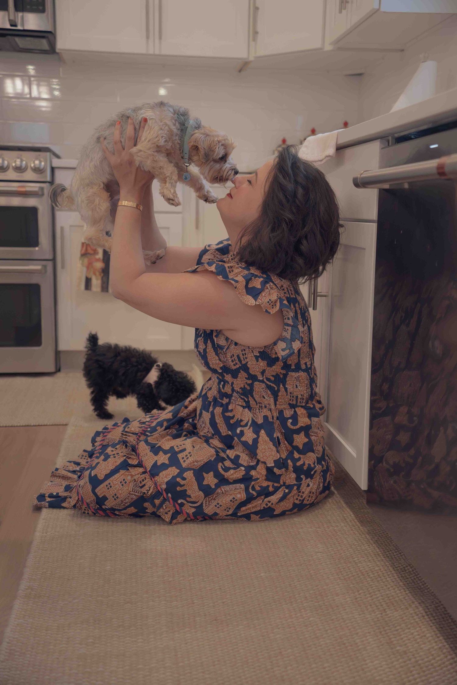 Woman in floral dress lifting small yorkie puppy up for a kiss in bright white kitchen.
