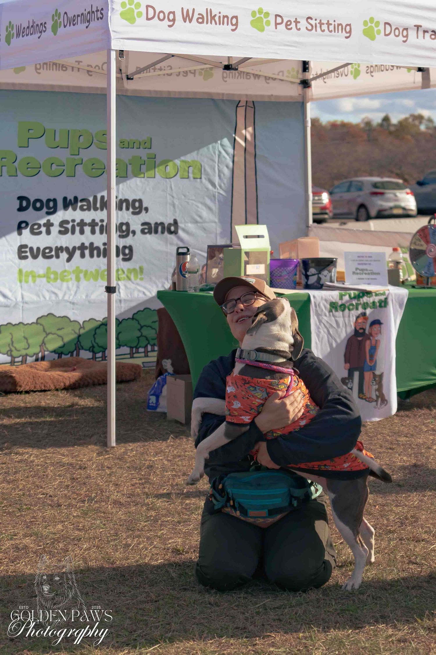 Person kneeling and hugging a dog at a Pups and Recreation pet services booth outdoors.