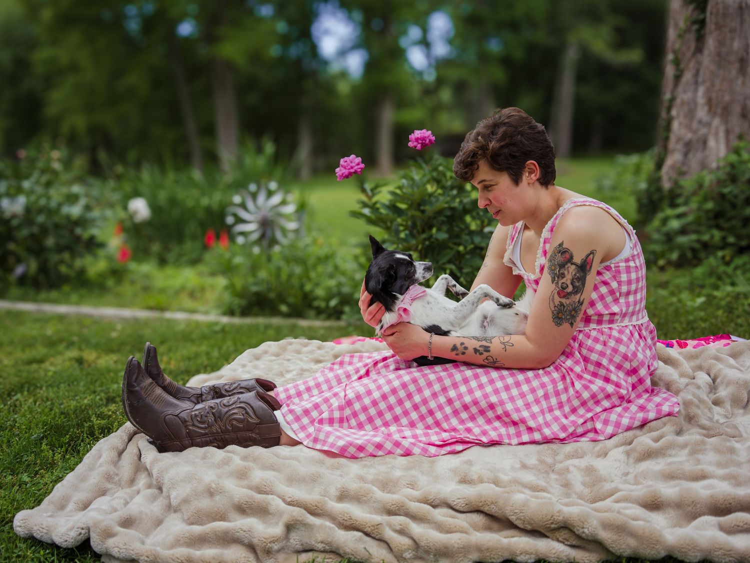 Woman in pink gingham dress and cowboy boots sits on blanket outdoors holding small dog in lush garden.