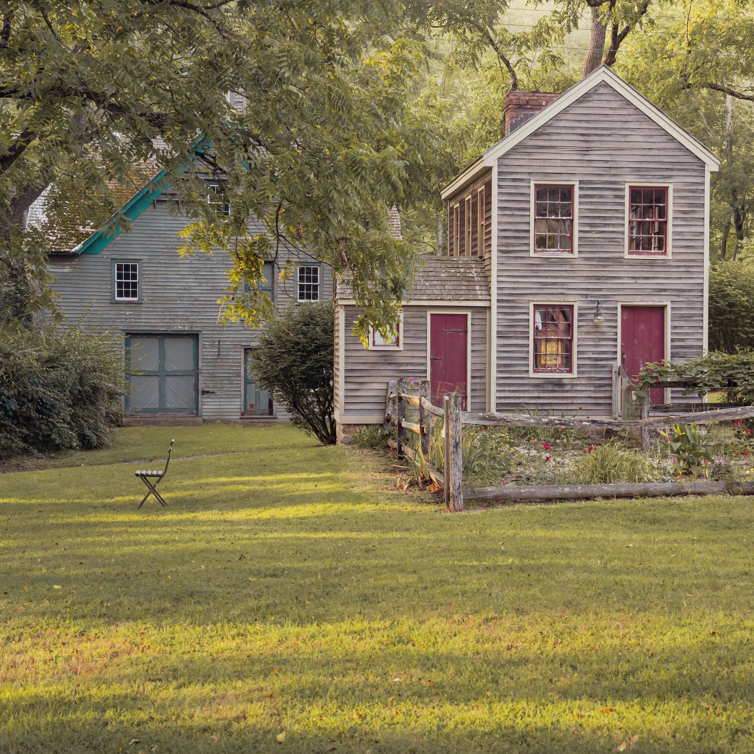 Rustic weathered wooden colonial farmhouse with red doors on green lawn under large shade trees.