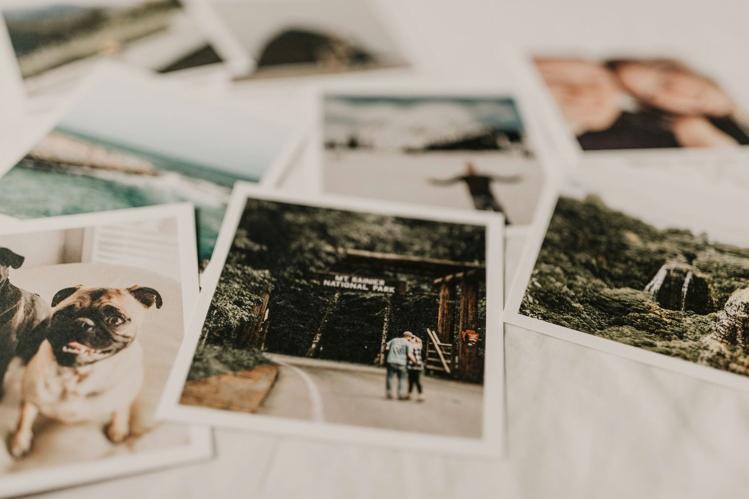 A collection of printed square photos scattered on a white surface, featuring a dog, landscapes, and a couple.