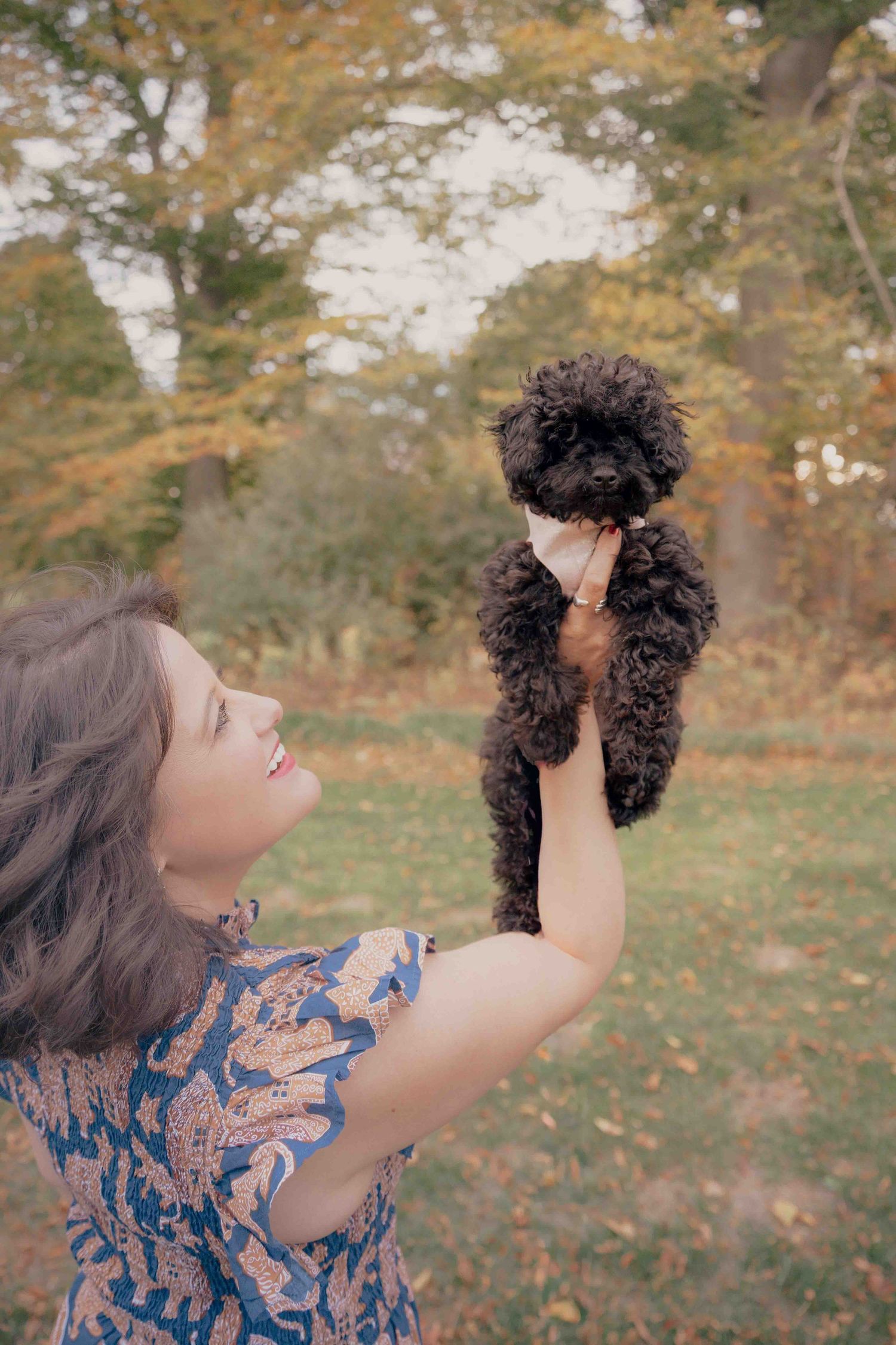 A smiling woman holds up a tiny black curly-haired puppy against a backdrop of autumn foliage.