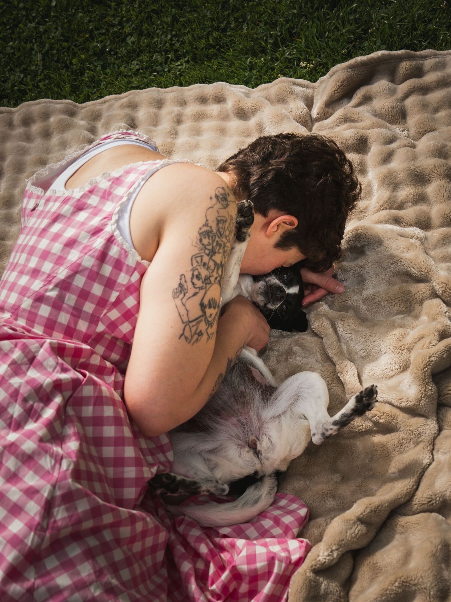 Person with tattoos in pink gingham dress cuddling a small gray dog on a blanket outdoors.