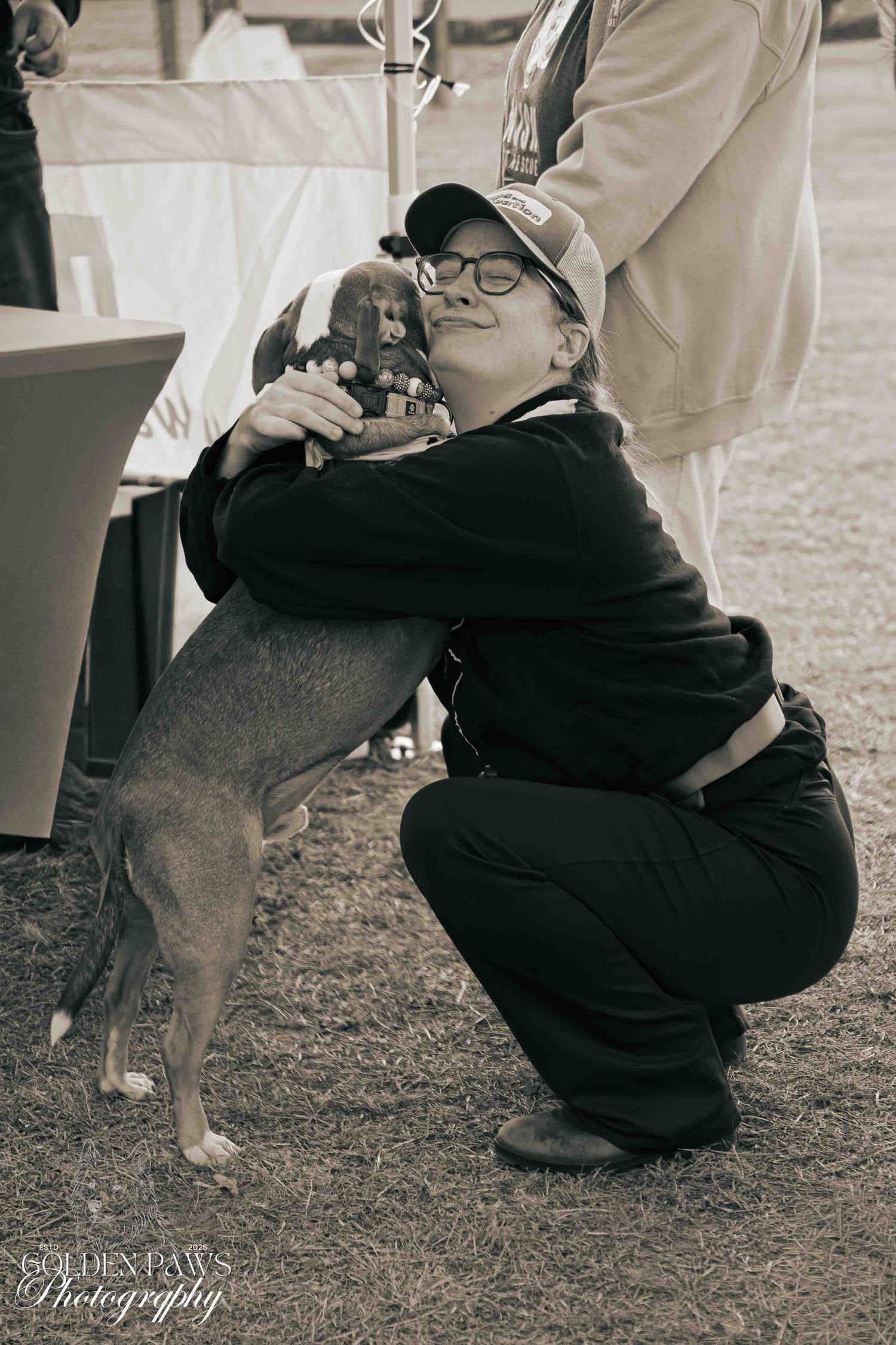 Woman in black outfit crouching and hugging a small dog at an outdoor pet event, black and white photo.