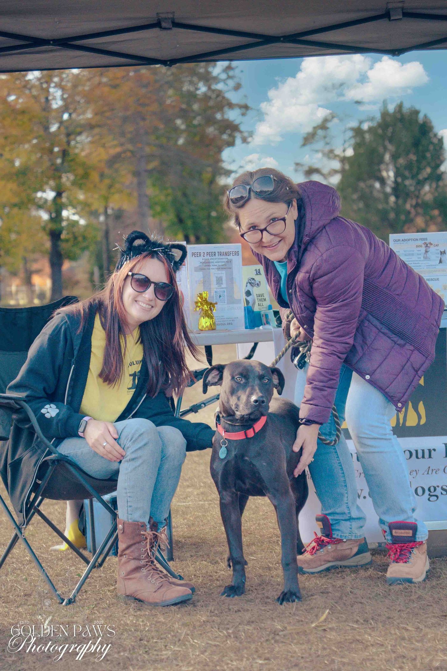 Two women pose with a black dog at an outdoor pet event under a canopy on a sunny autumn day.