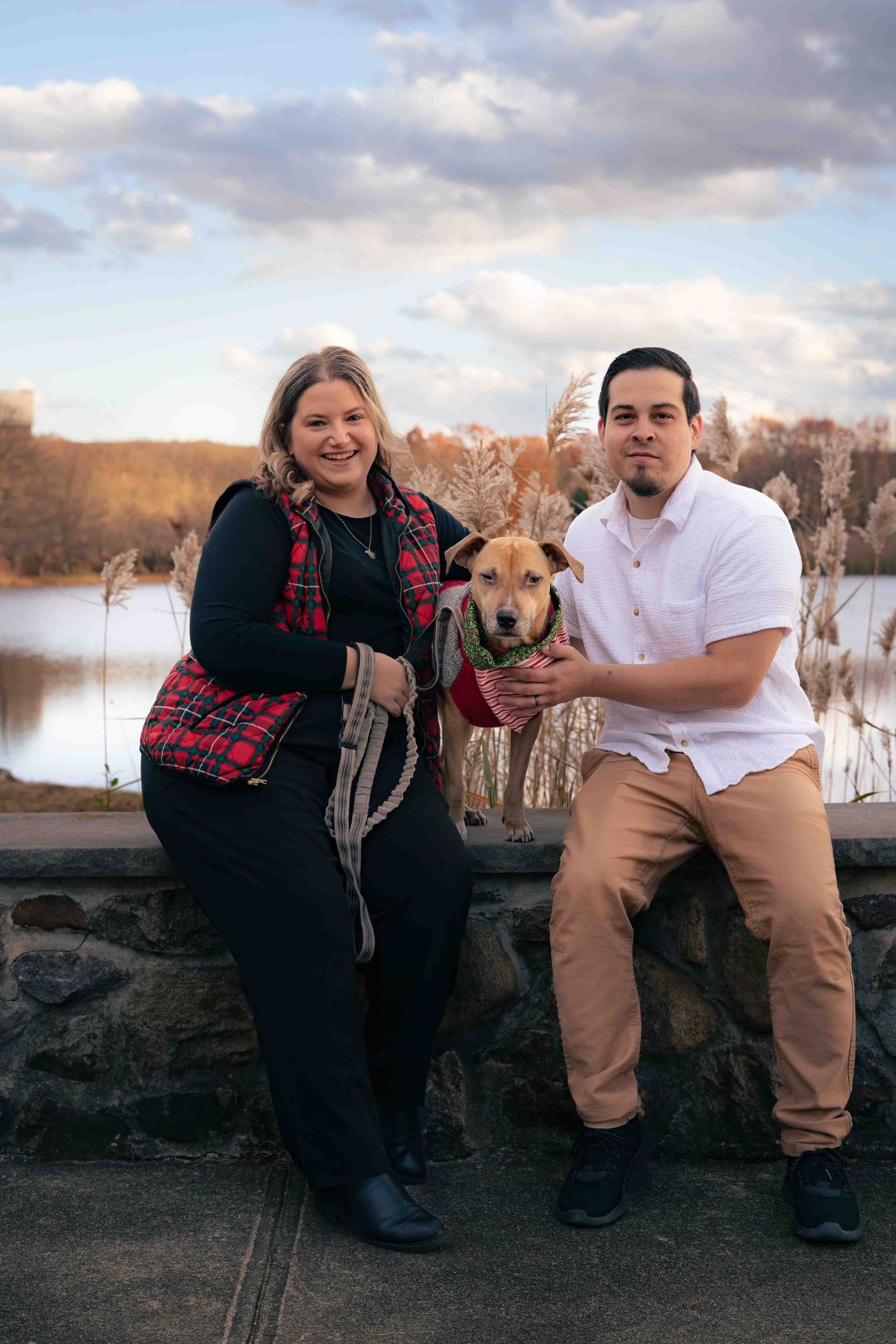 Happy couple holding their dog by a lakeside with dramatic cloudy skies and autumn scenery.