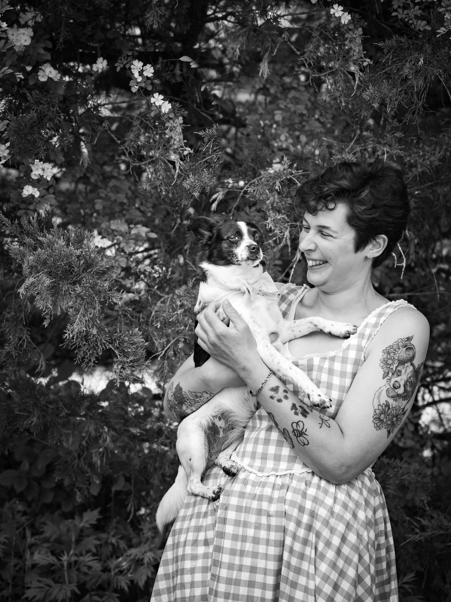 Black and white photo of laughing woman holding small dog in gingham dress outdoors.