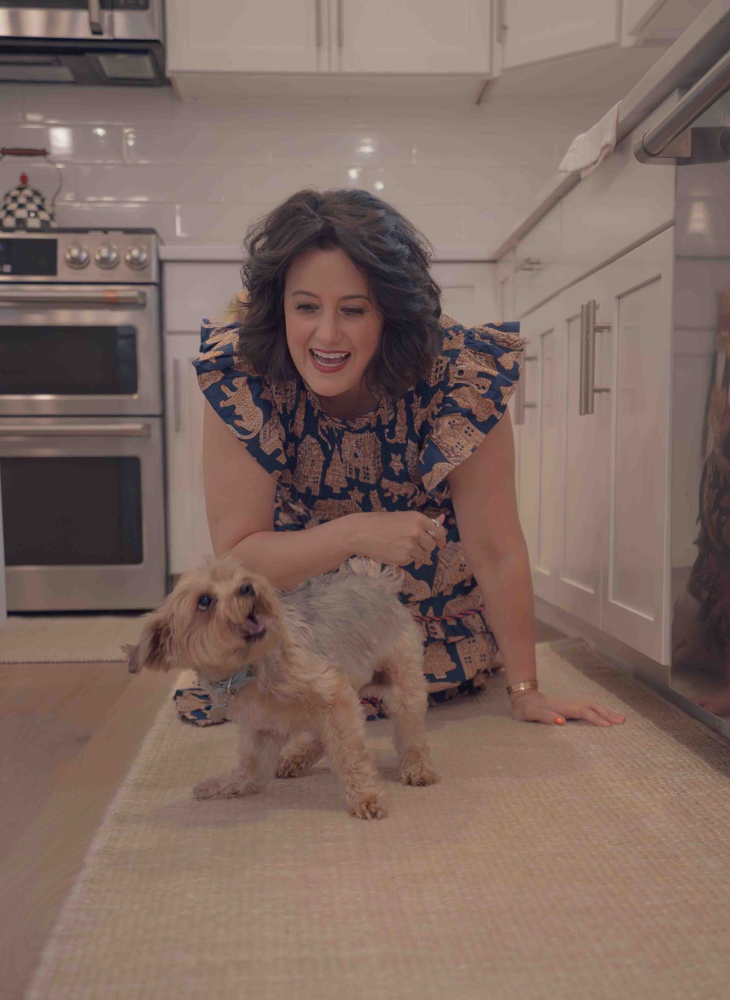 Happy woman in blue floral dress bending down to pet a small yorkie dog in modern white kitchen.