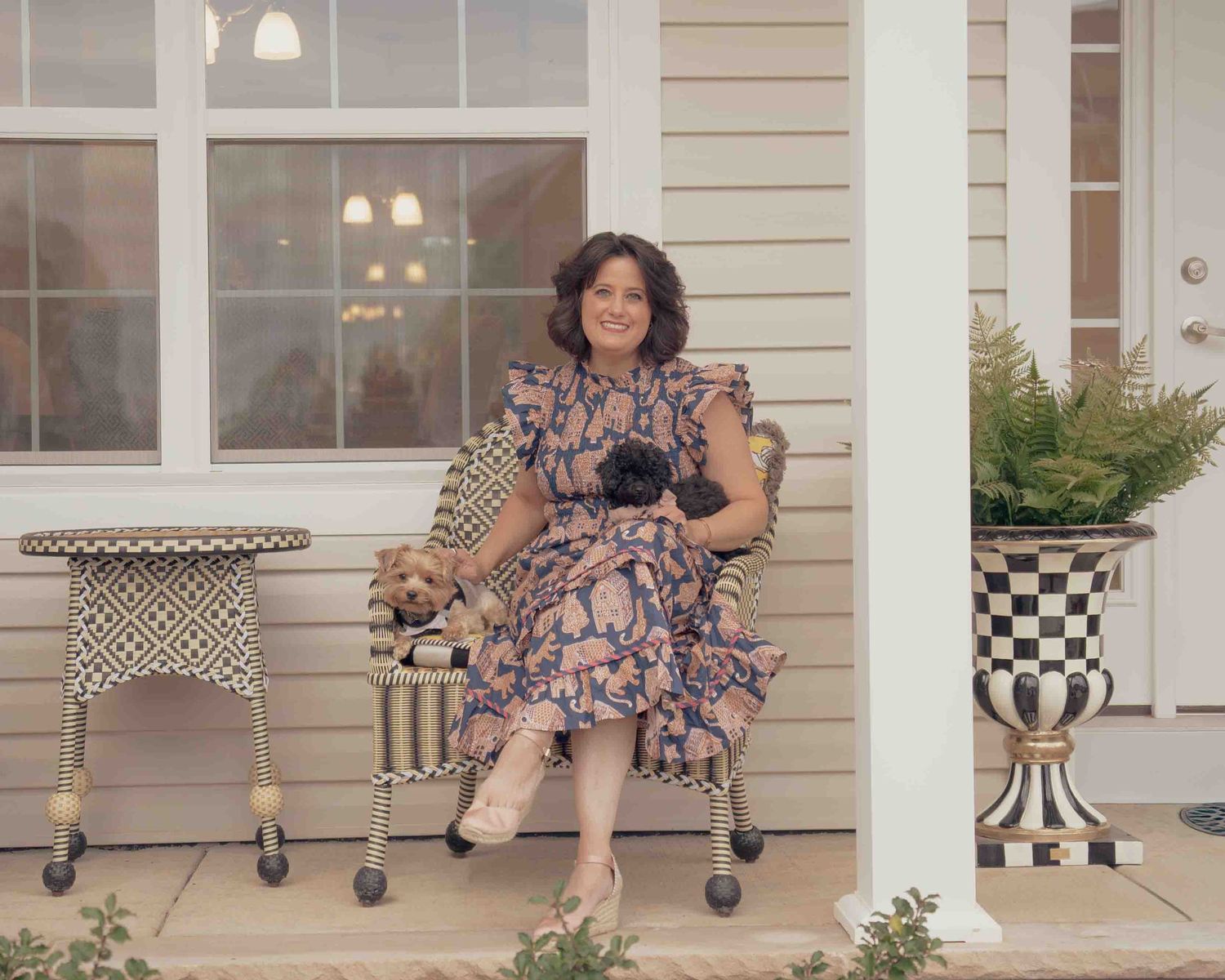 Smiling woman in floral dress sitting on porch bench holding two small dogs with checkered decor.