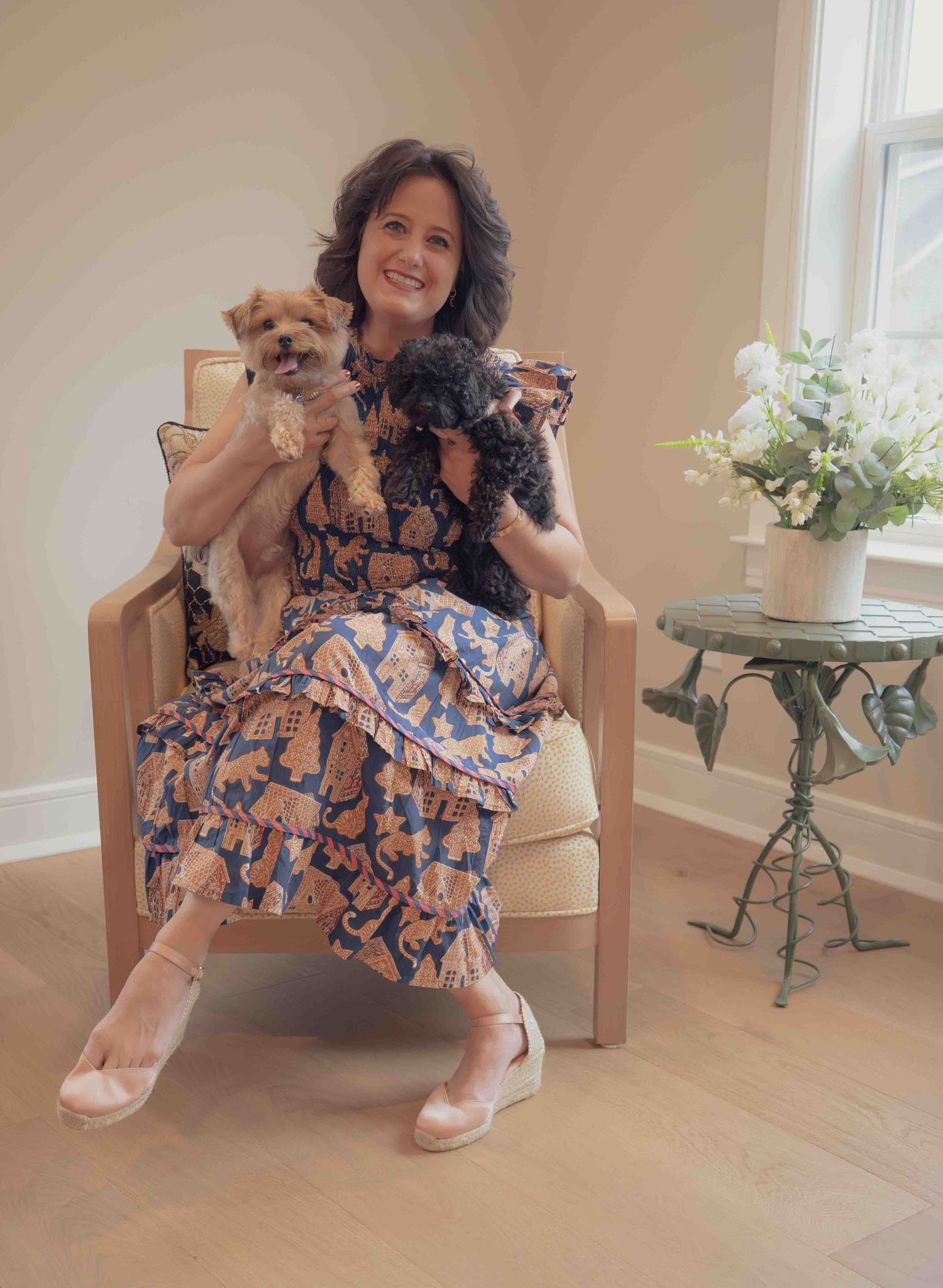 Smiling woman in blue floral dress seated in chair holding yorkie and black poodle puppies indoors.