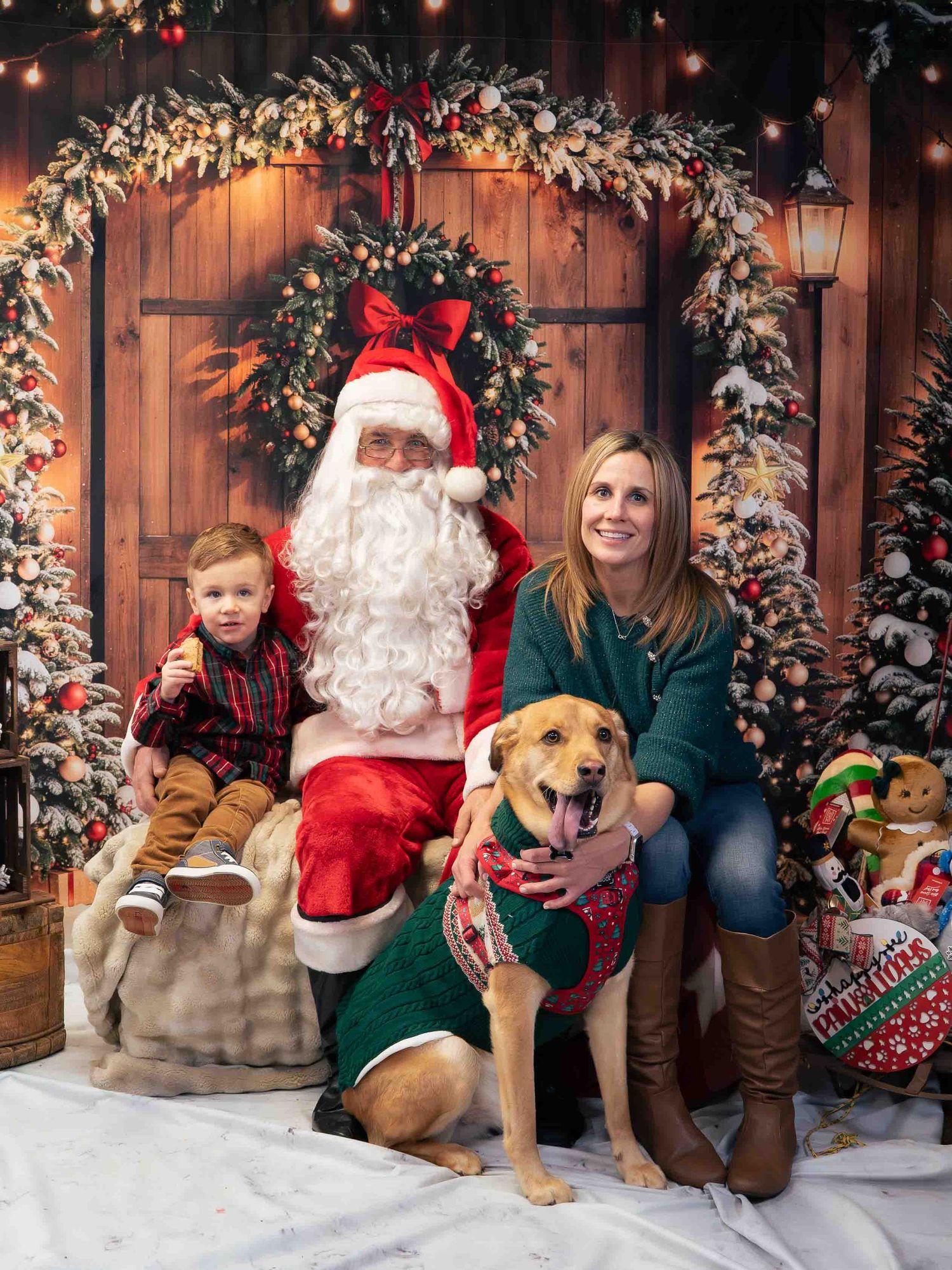 Family Christmas photo with Santa, a young boy, woman, and golden dog in festive sweater against holiday backdrop.