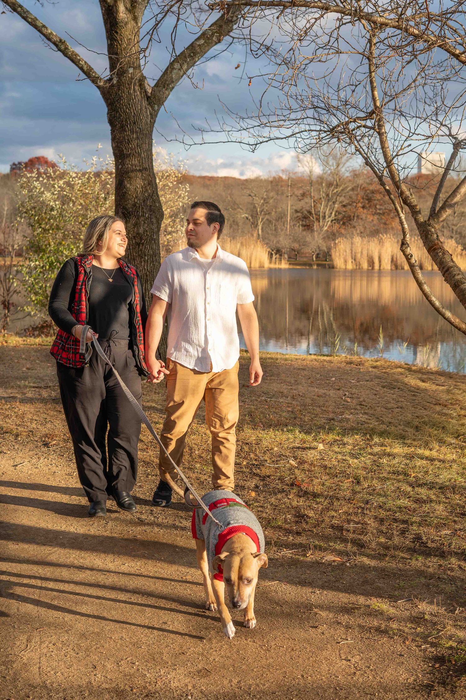 Couple holding hands while walking tan dog in red sweater along sunlit lakeside path.