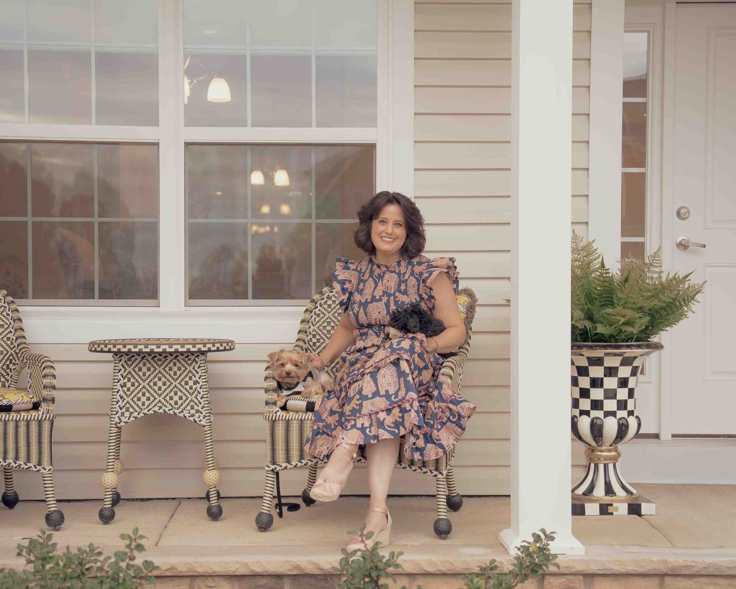 A smiling woman in a floral dress sits on a porch with checkered decor holding a small black dog.