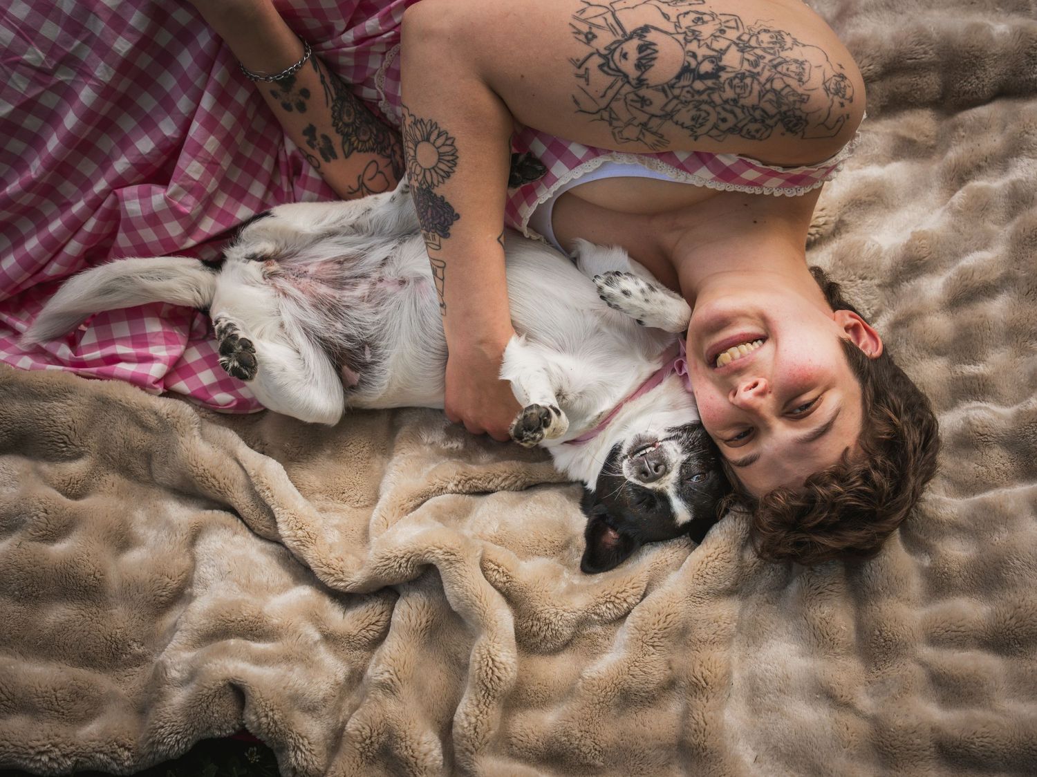 Smiling tattooed woman lying on a fluffy beige blanket cuddling with an adorable black and white dog.