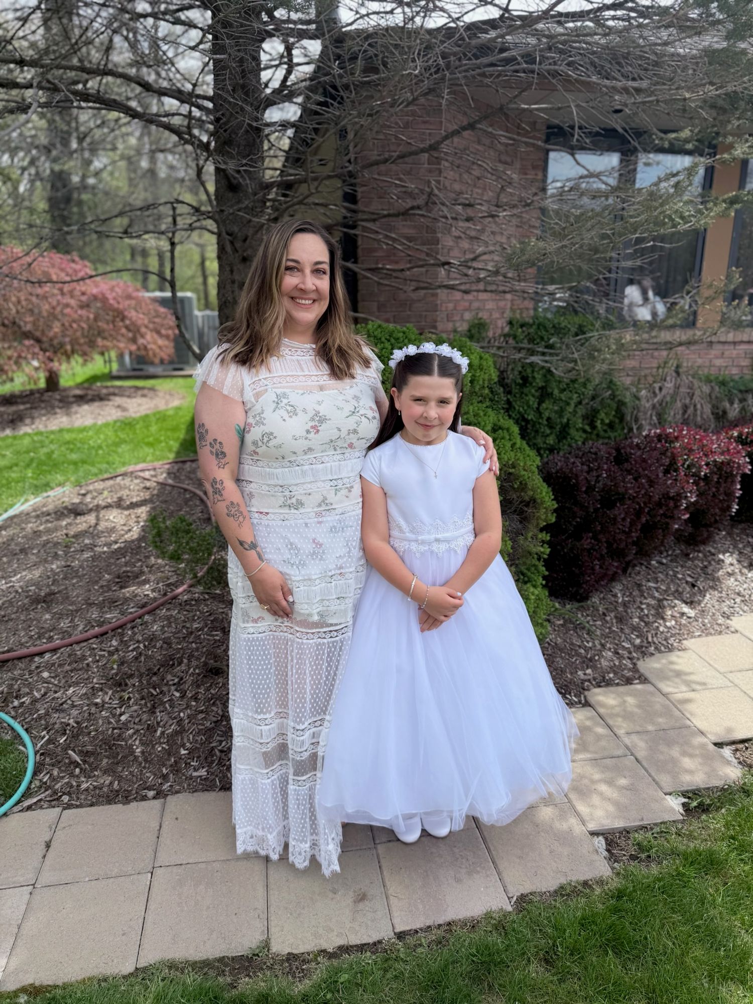 A woman in a lace dress stands beside a young girl in a white First Communion gown outdoors.