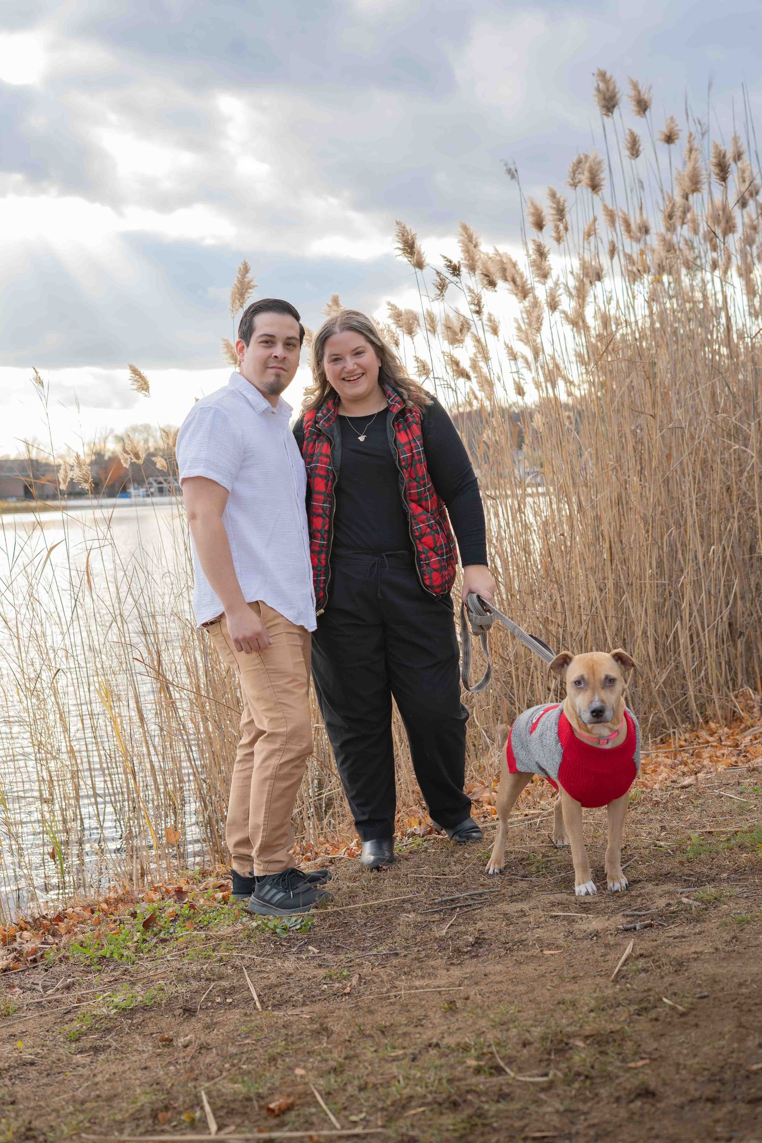 Couple posing with tan dog in red sweater near lake with dramatic cloudy sky.
