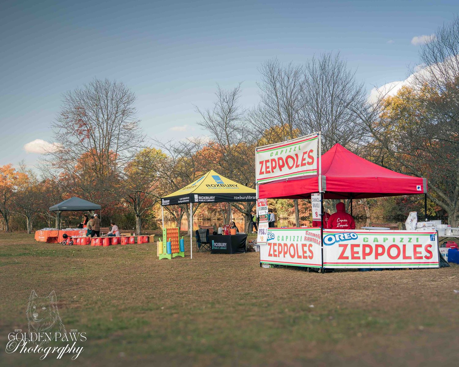 Outdoor zeppoles food vendor tents at a fall festival in a park with colorful foliage and open green space.