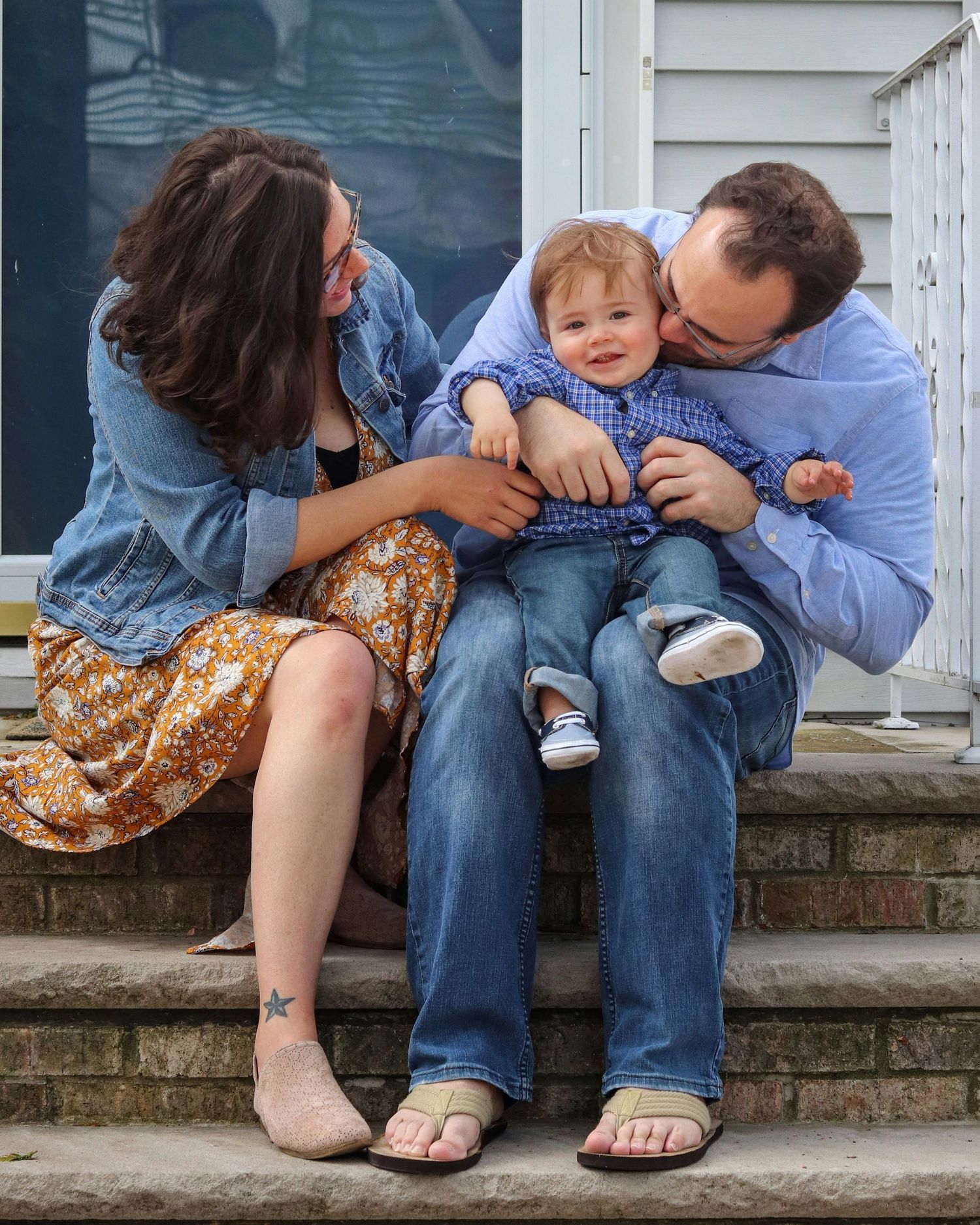 A happy family with a baby boy sitting on porch steps, parents leaning in to kiss and cuddle their smiling toddler.