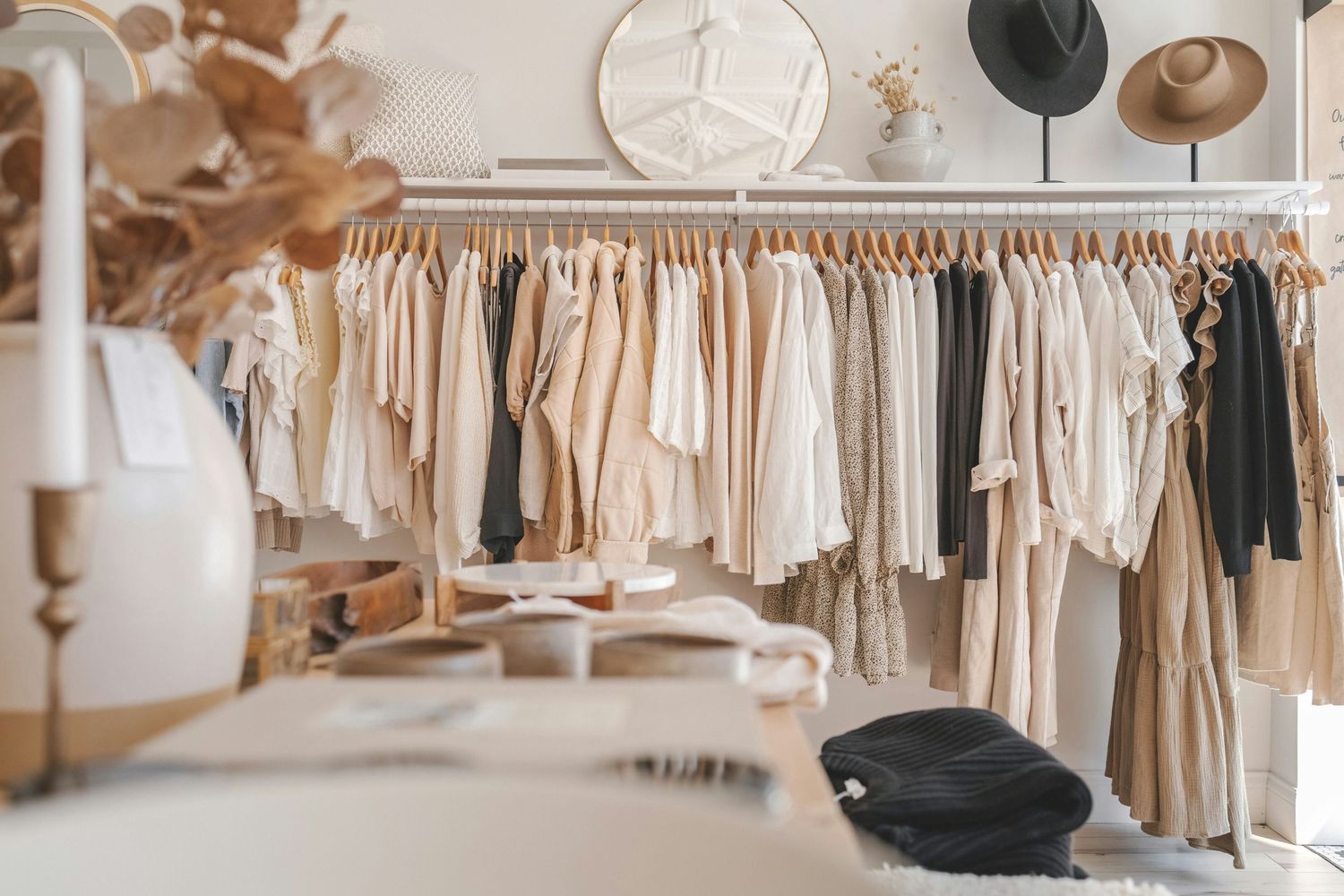 Boutique clothing rack displaying neutral-toned garments in beige, white, and black, with hats and decor in a stylish shop.