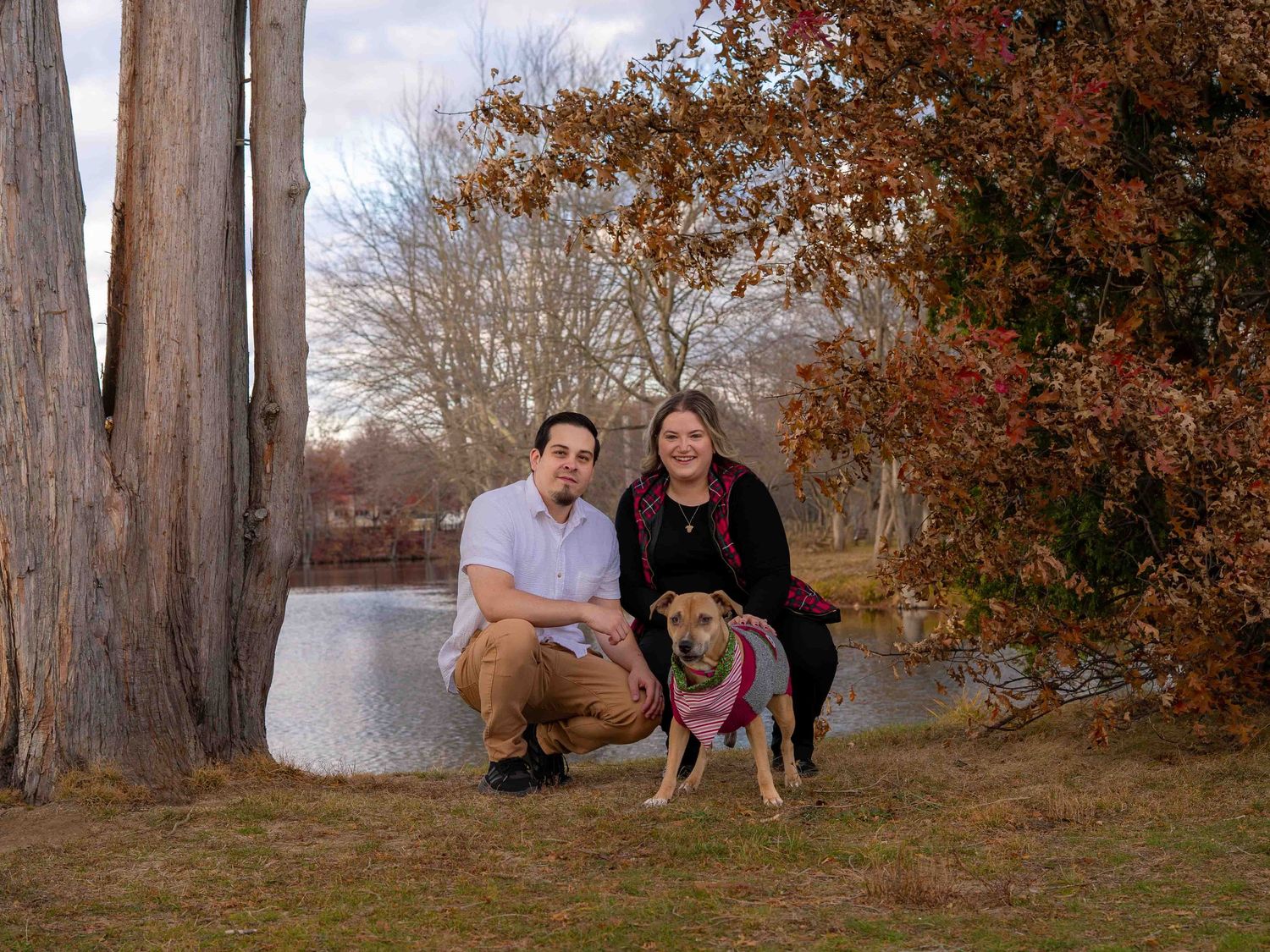 Couple crouching with their dog on a leash near a pond surrounded by fall foliage and tall trees.