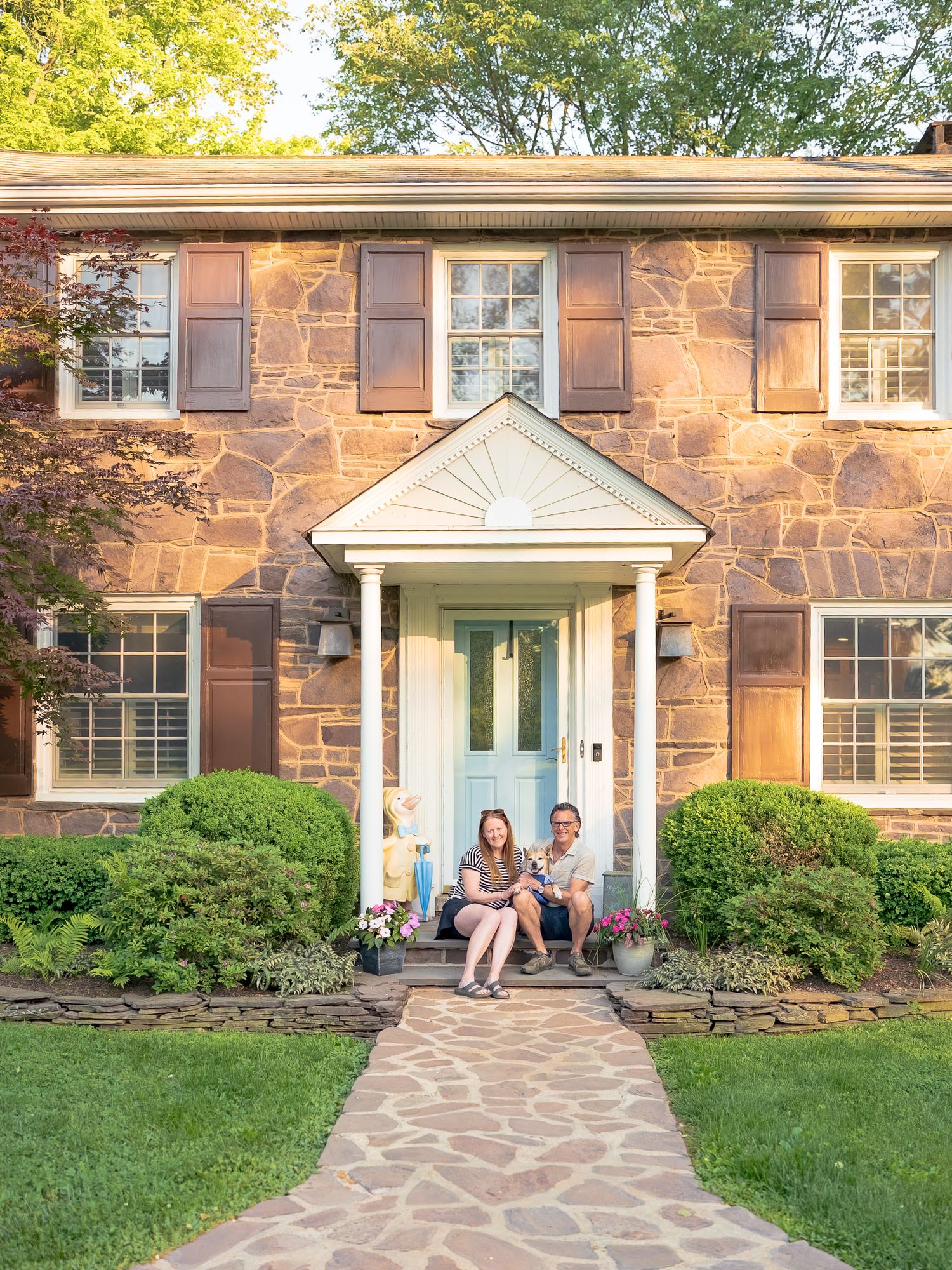 Couple sitting on the front porch of a charming two-story stone colonial home with blue door and lush landscaping.