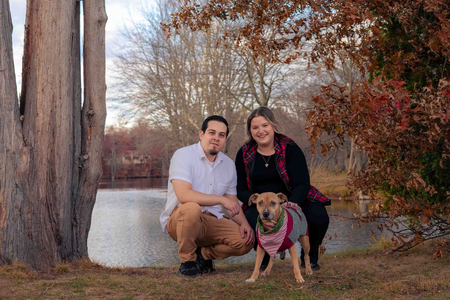 Couple crouching with tan dog near pond surrounded by autumn trees.