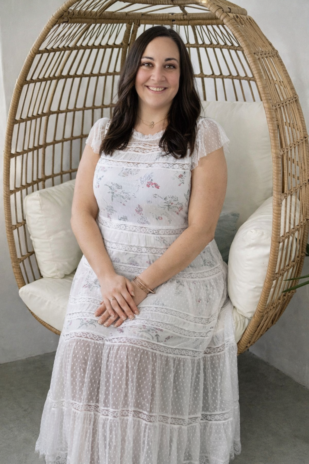 Smiling brunette woman in white floral lace dress sitting in a wicker egg chair.