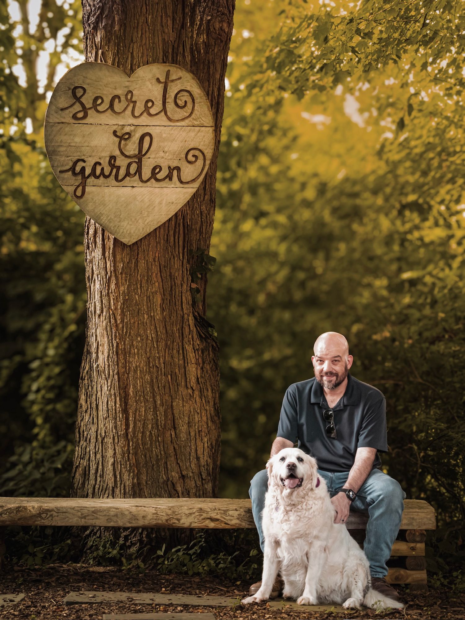 A bald man sits on a bench beside a white Golden Retriever under a Secret Garden tree sign.