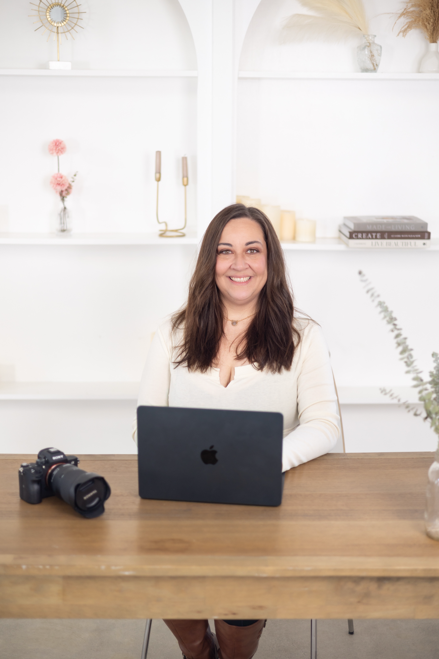 Smiling brunette woman sits at wooden desk with Apple laptop and camera in bright white office.