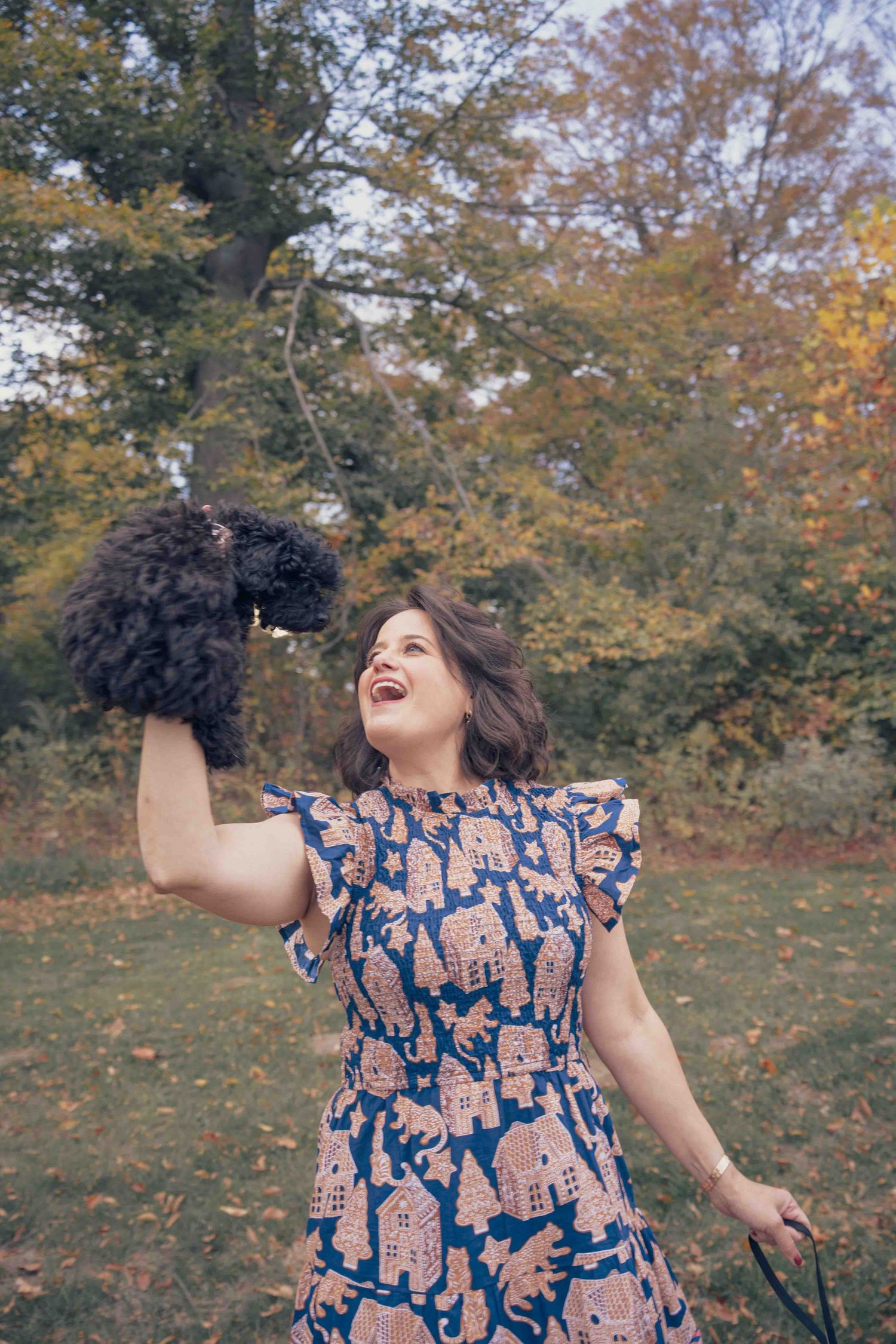 A joyful woman in a floral dress lifts a small black curly-haired dog in an autumn park setting.