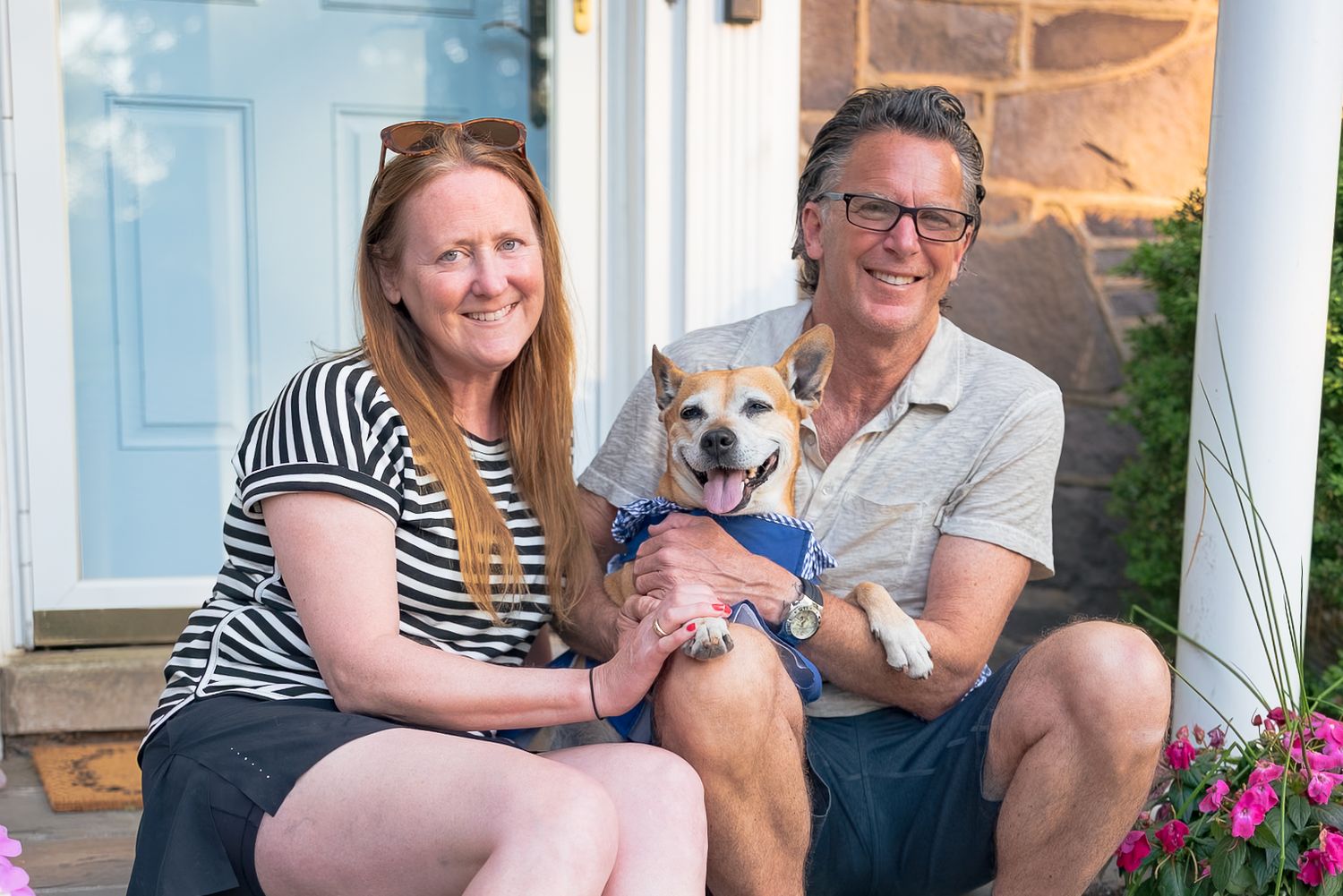 Smiling couple sitting on front porch steps holding a Shiba Inu dog outside a white home with stone accents.