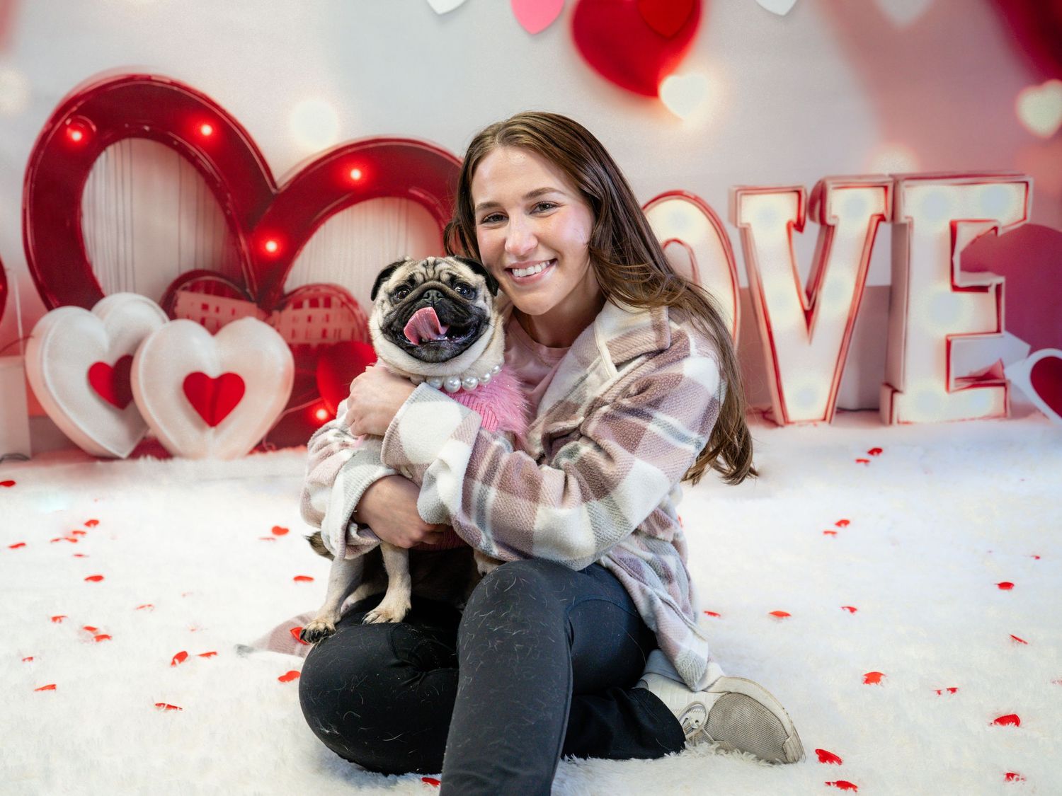 Young woman smiling and holding a pug dog in front of a Valentine's Day backdrop with hearts and LOVE letters.