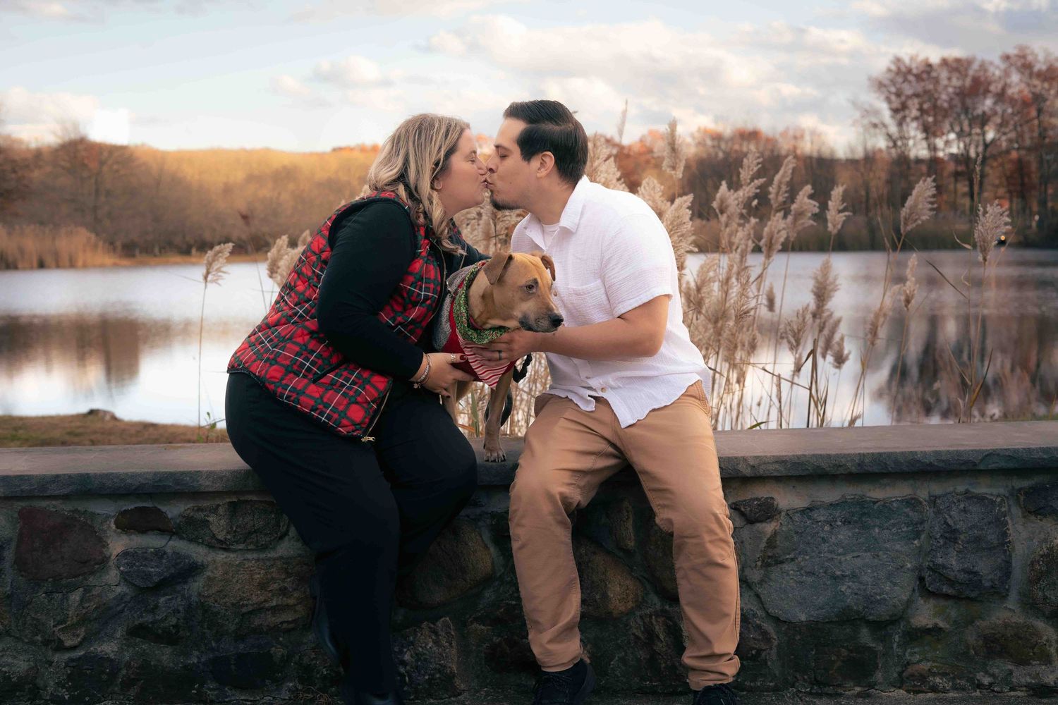 Couple sharing a kiss while holding their dog near a serene lake during autumn season.