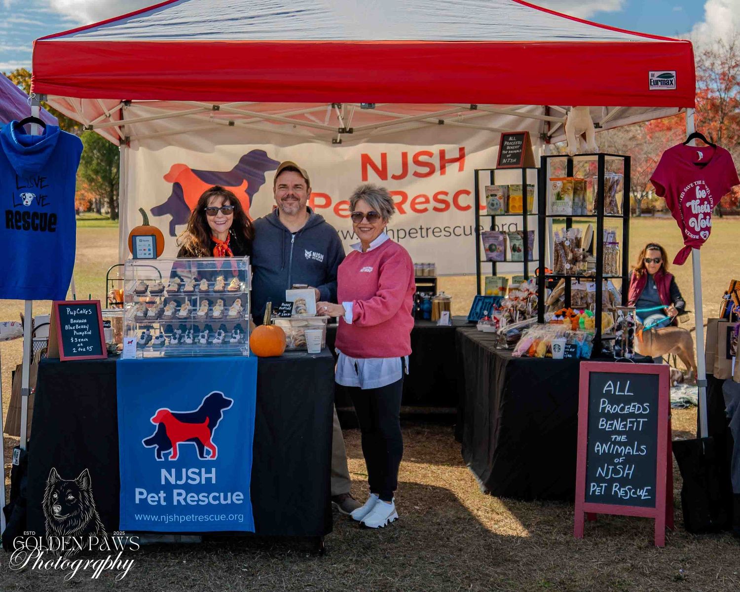 Three volunteers smile at an NJSH Pet Rescue outdoor fundraiser booth displaying merchandise and animal adoption information.