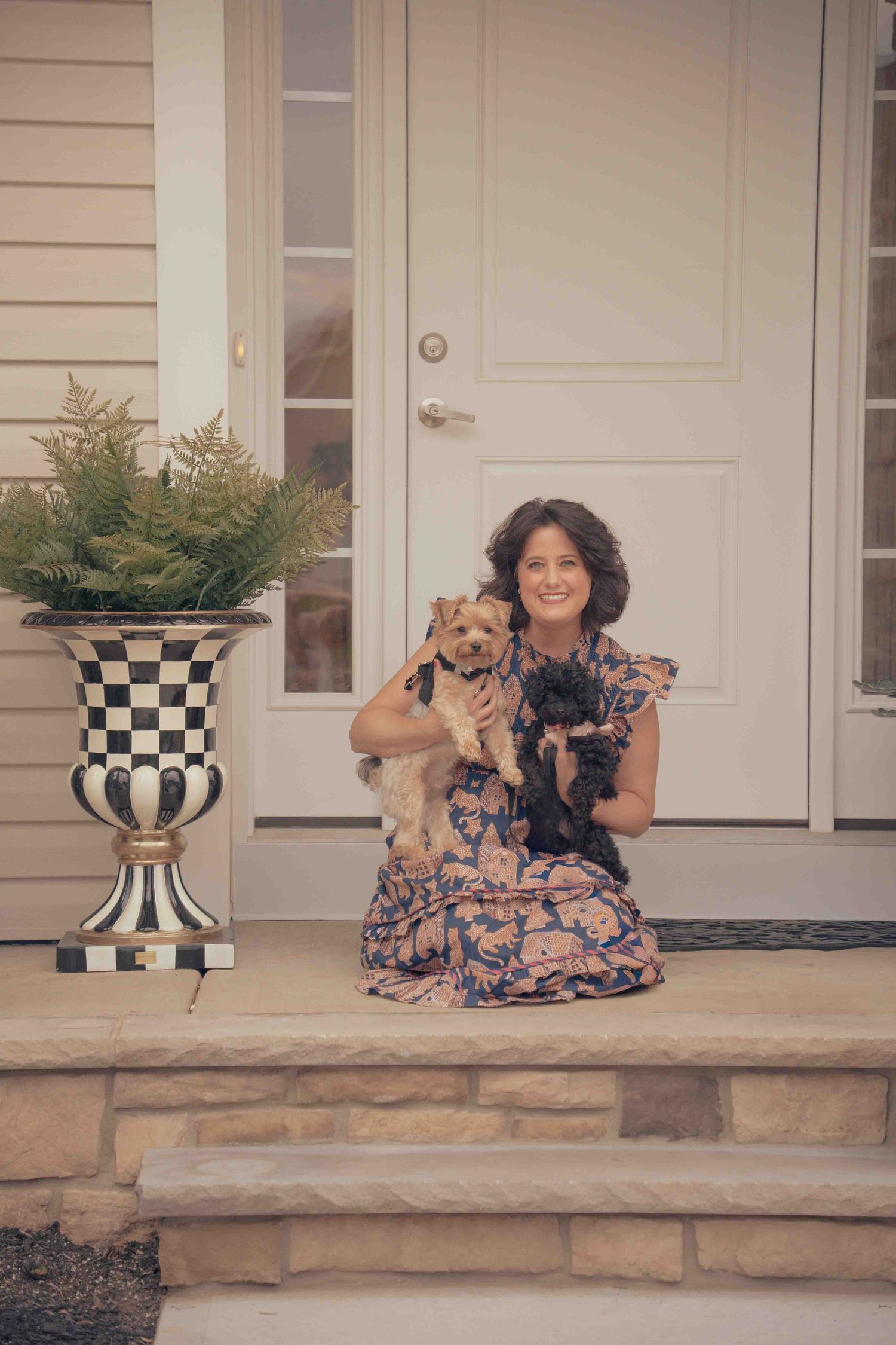 Smiling woman in floral dress kneeling on front porch steps holding two small dogs.