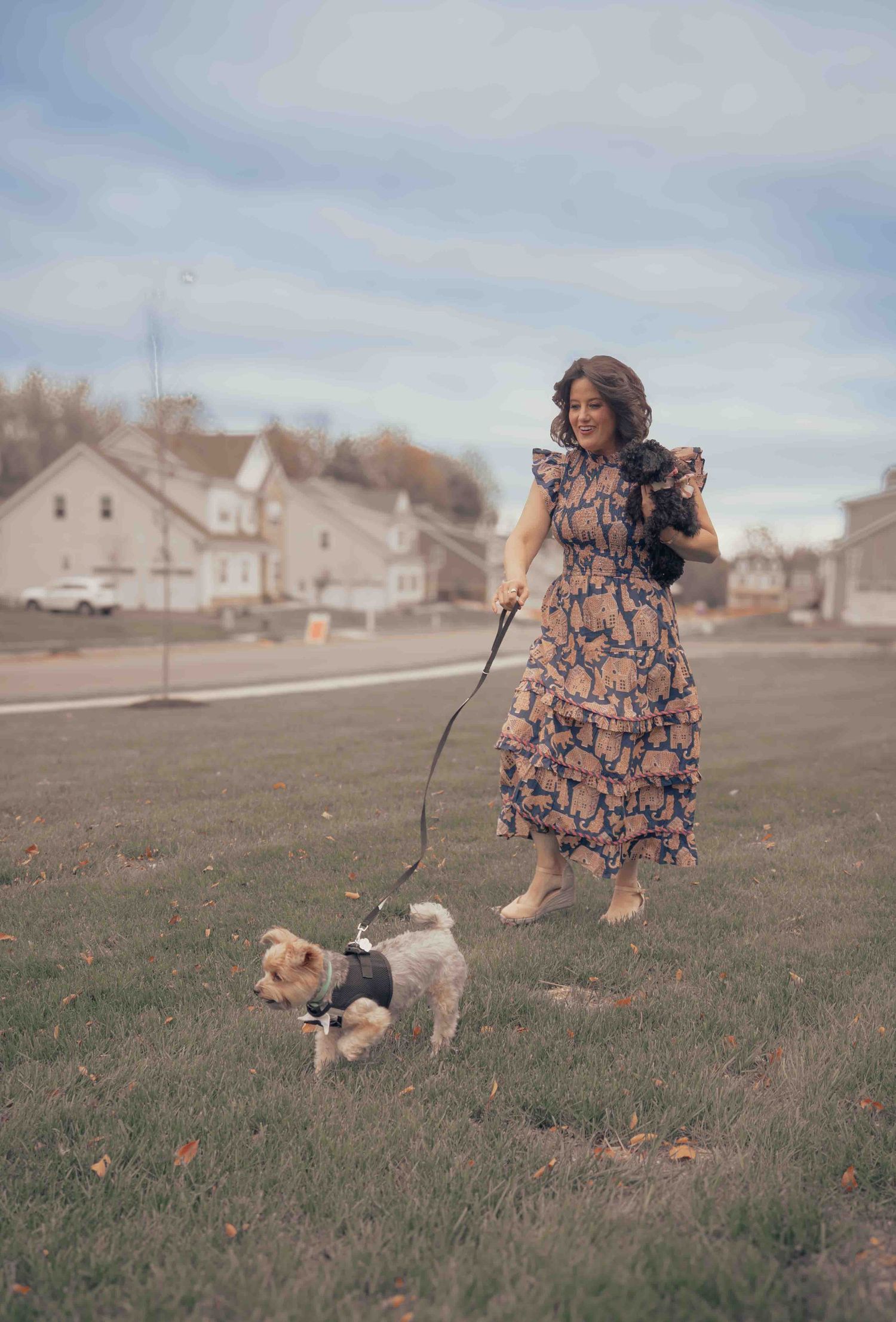 A woman in a floral midi dress walks a small Yorkshire Terrier on a leash across a green lawn.