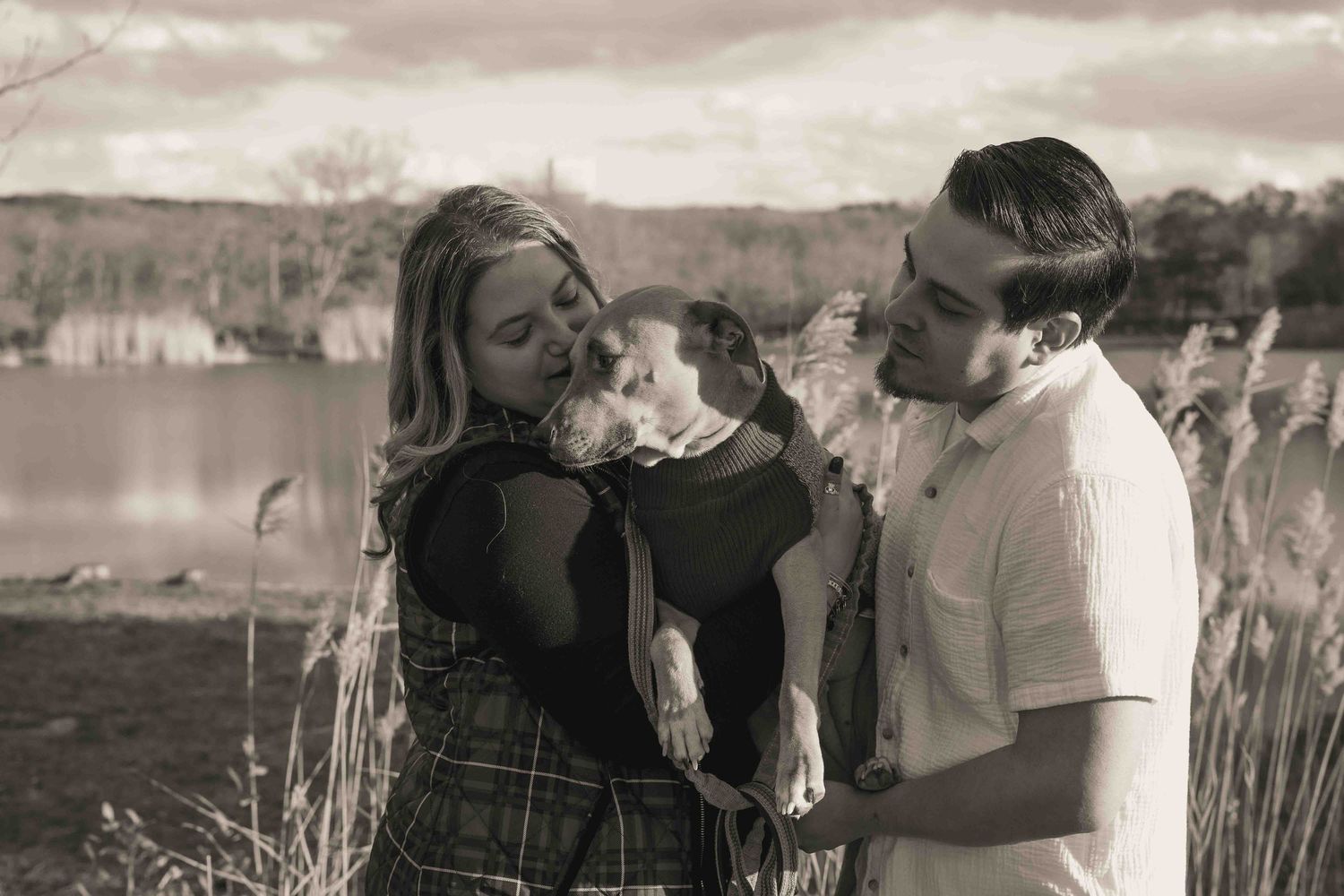 Black and white photo of a couple lovingly holding their dog near a lake with tall grass.