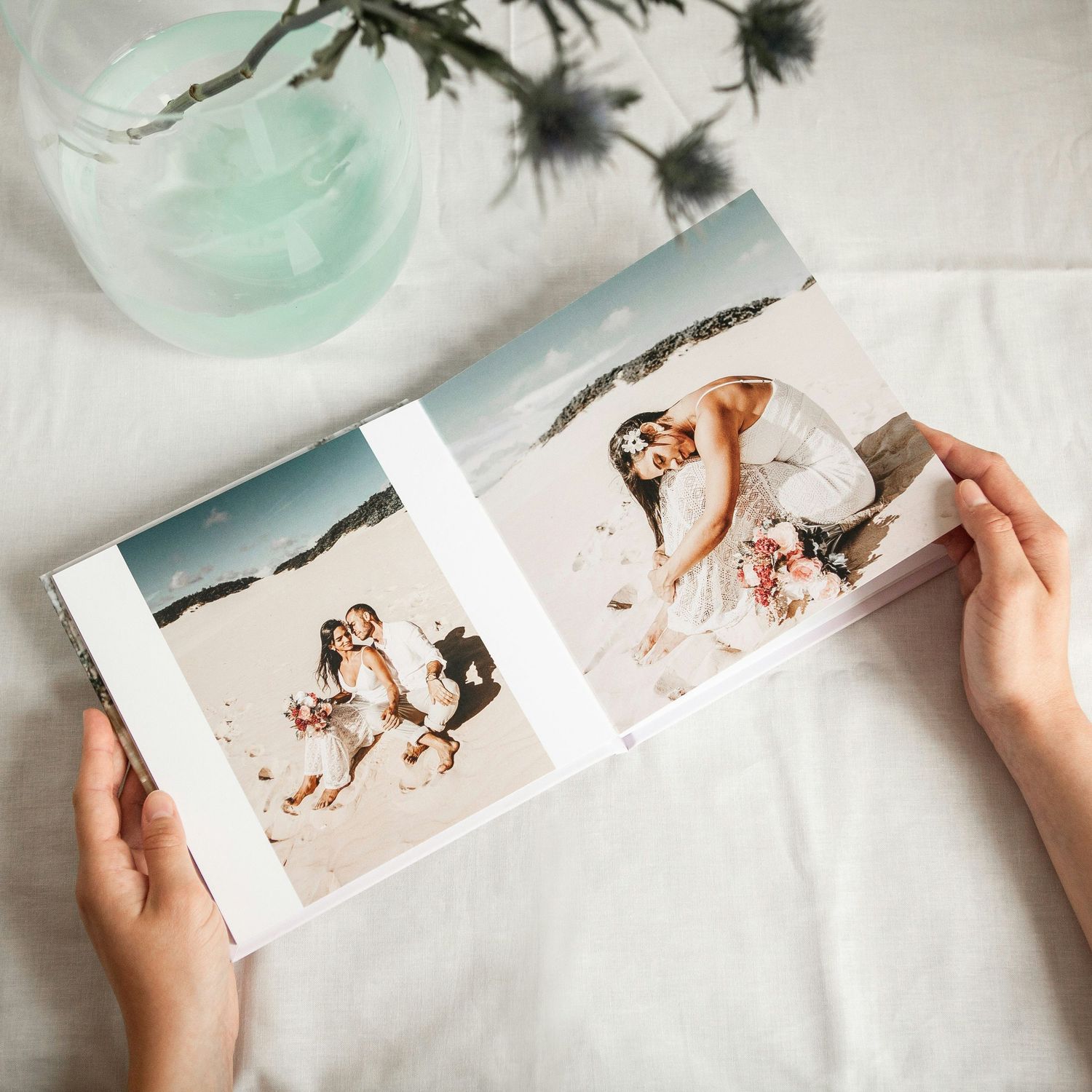 Hands holding open a wedding photo album with beach couple photos, displayed on a white linen surface with flowers.
