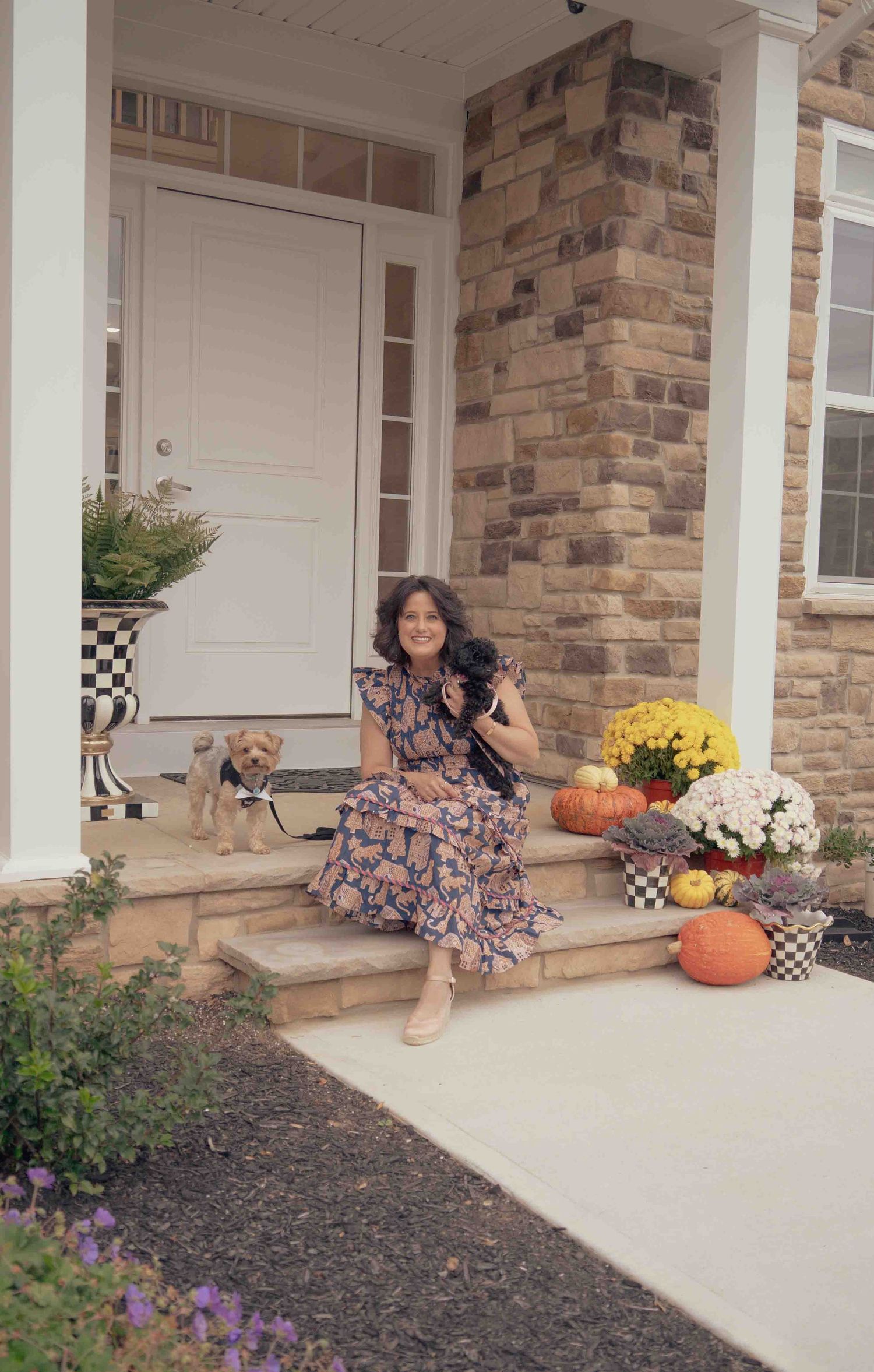 Woman in floral dress sitting on stone front porch steps with yorkie dog and fall decorations.