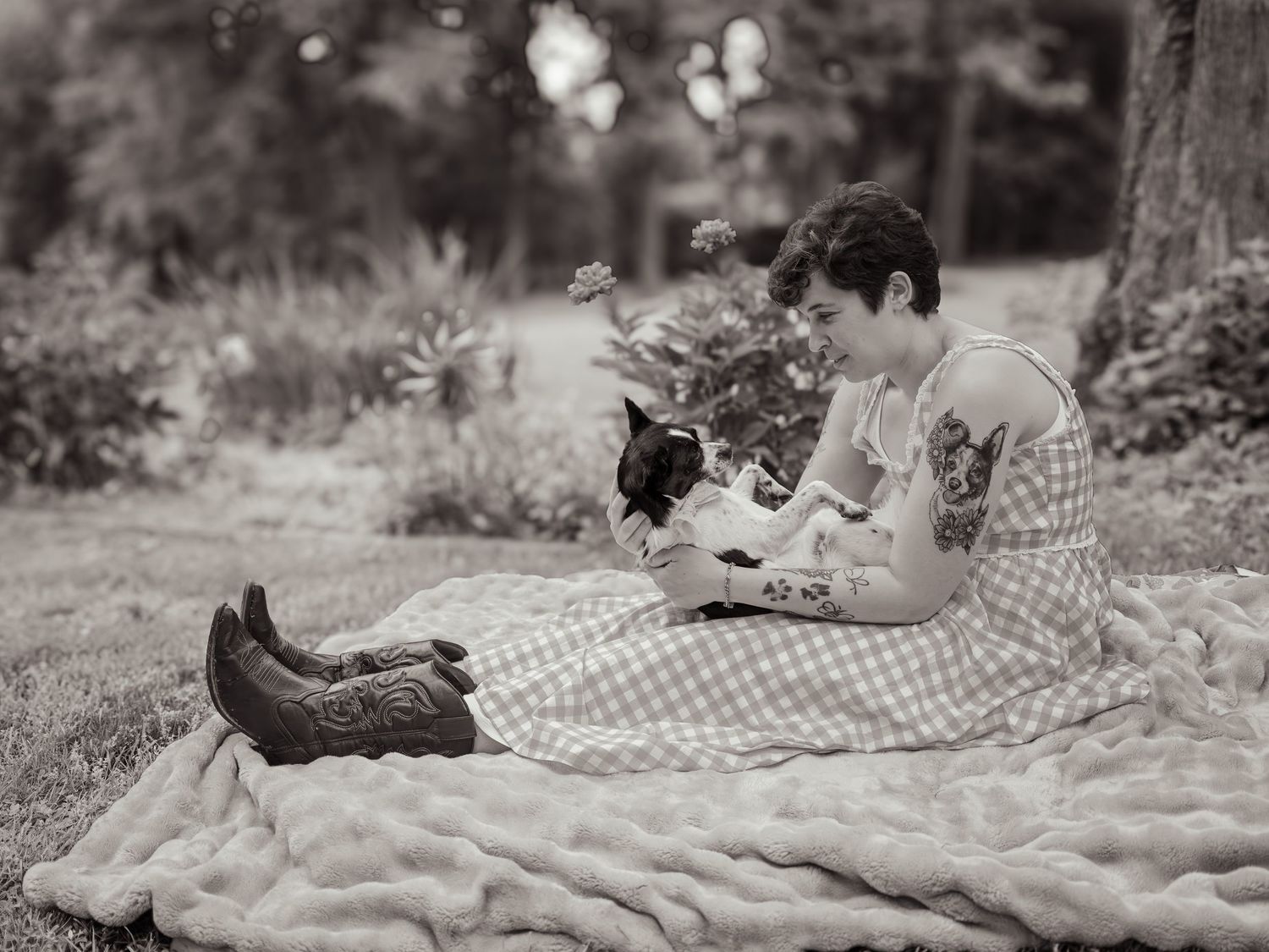 Woman in gingham dress sits on blanket outdoors, holding a cat, with cowboy boots nearby, black and white photo.