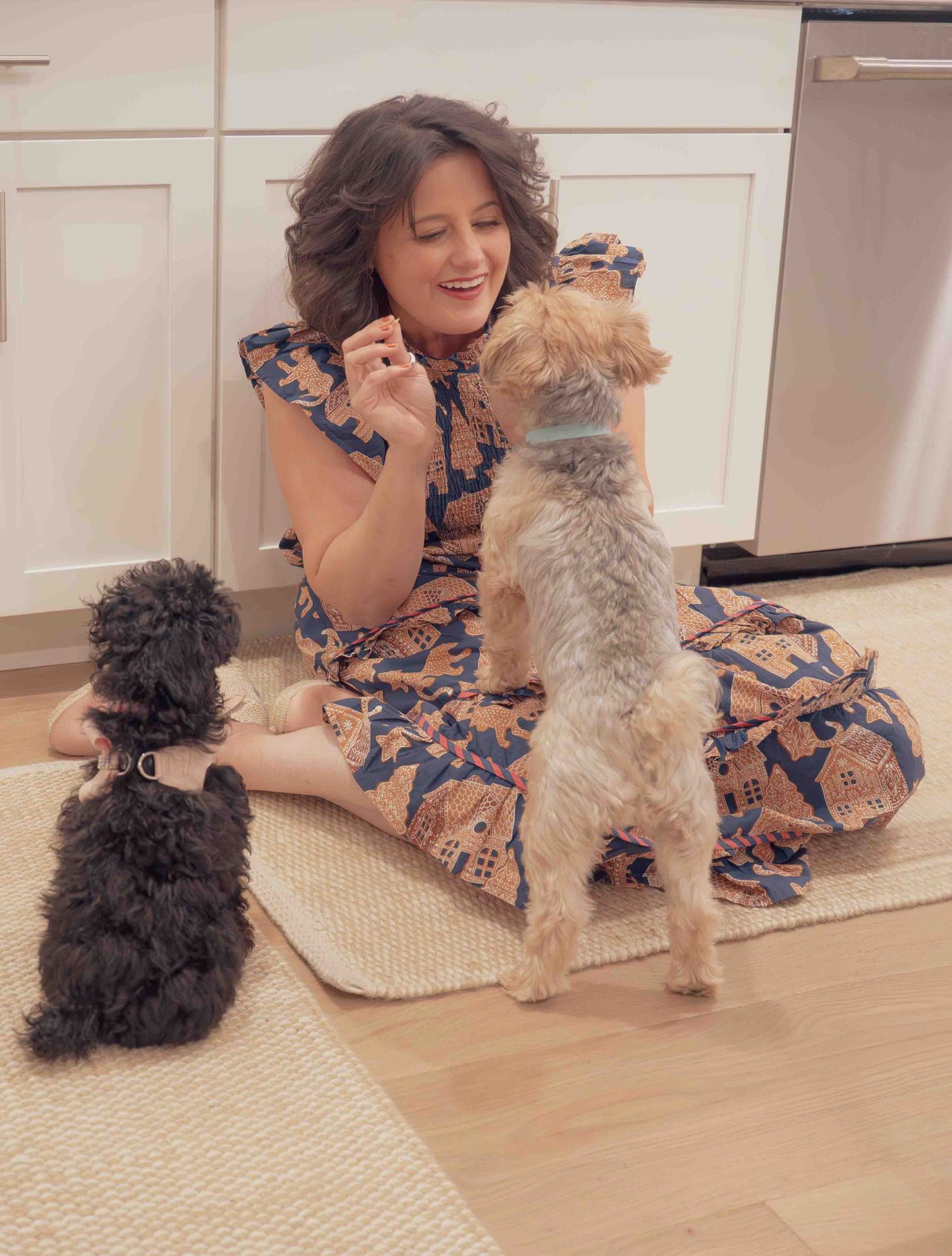 Woman in floral dress sitting on kitchen floor playing with two small dogs.