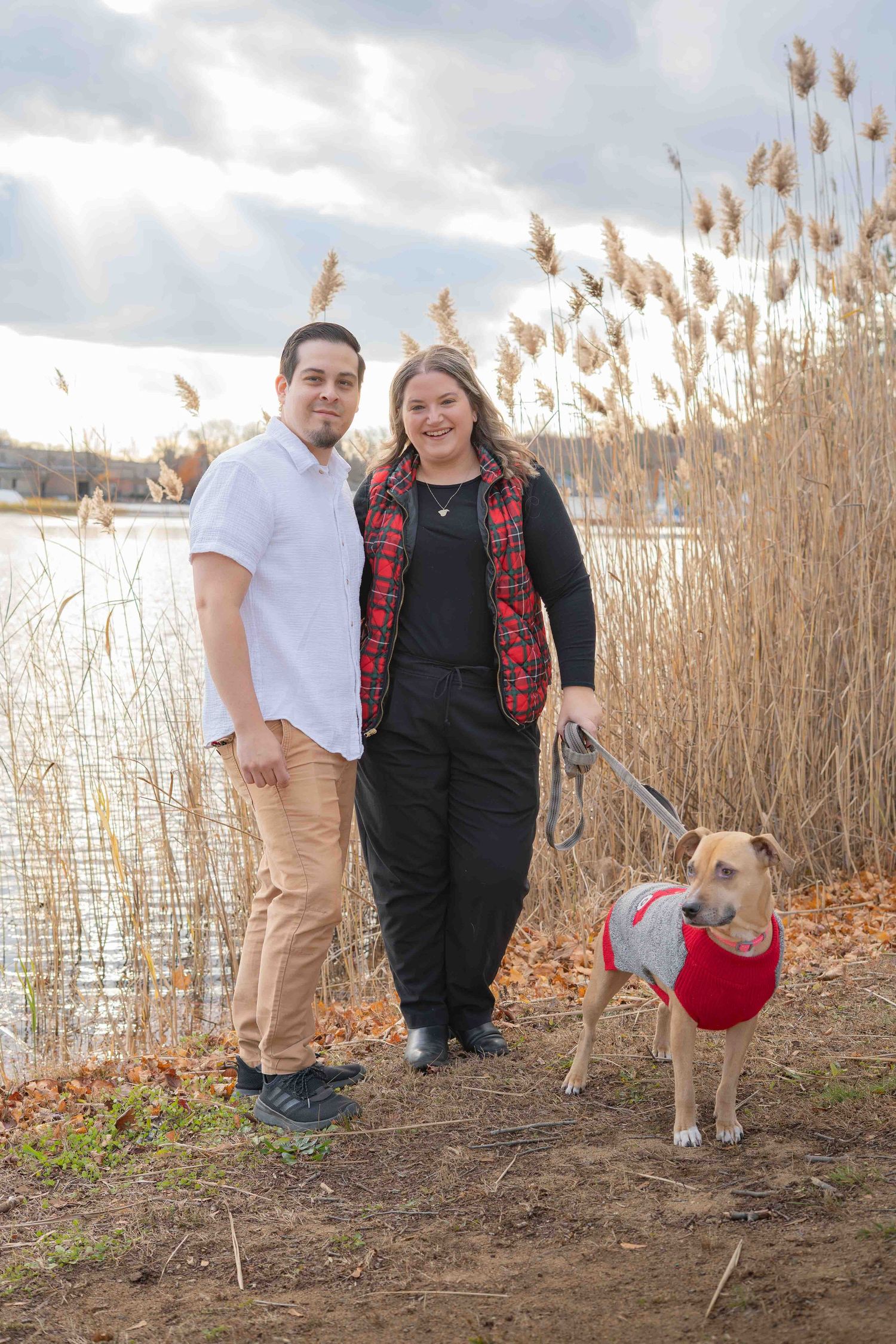 Couple standing by lakeside reeds with tan dog in red sweater on leash.