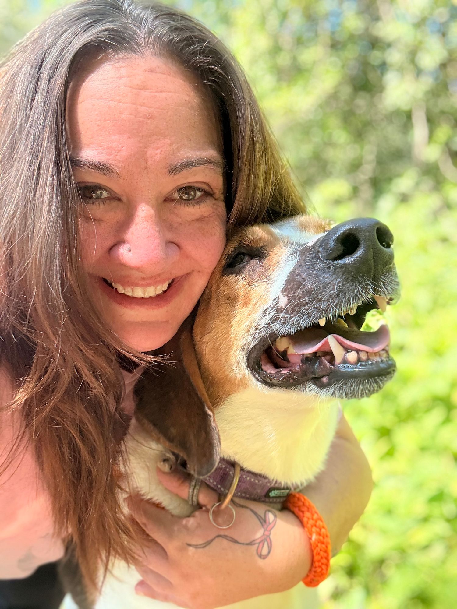 A smiling woman takes a sunny outdoor selfie with her tan and black dog in nature.