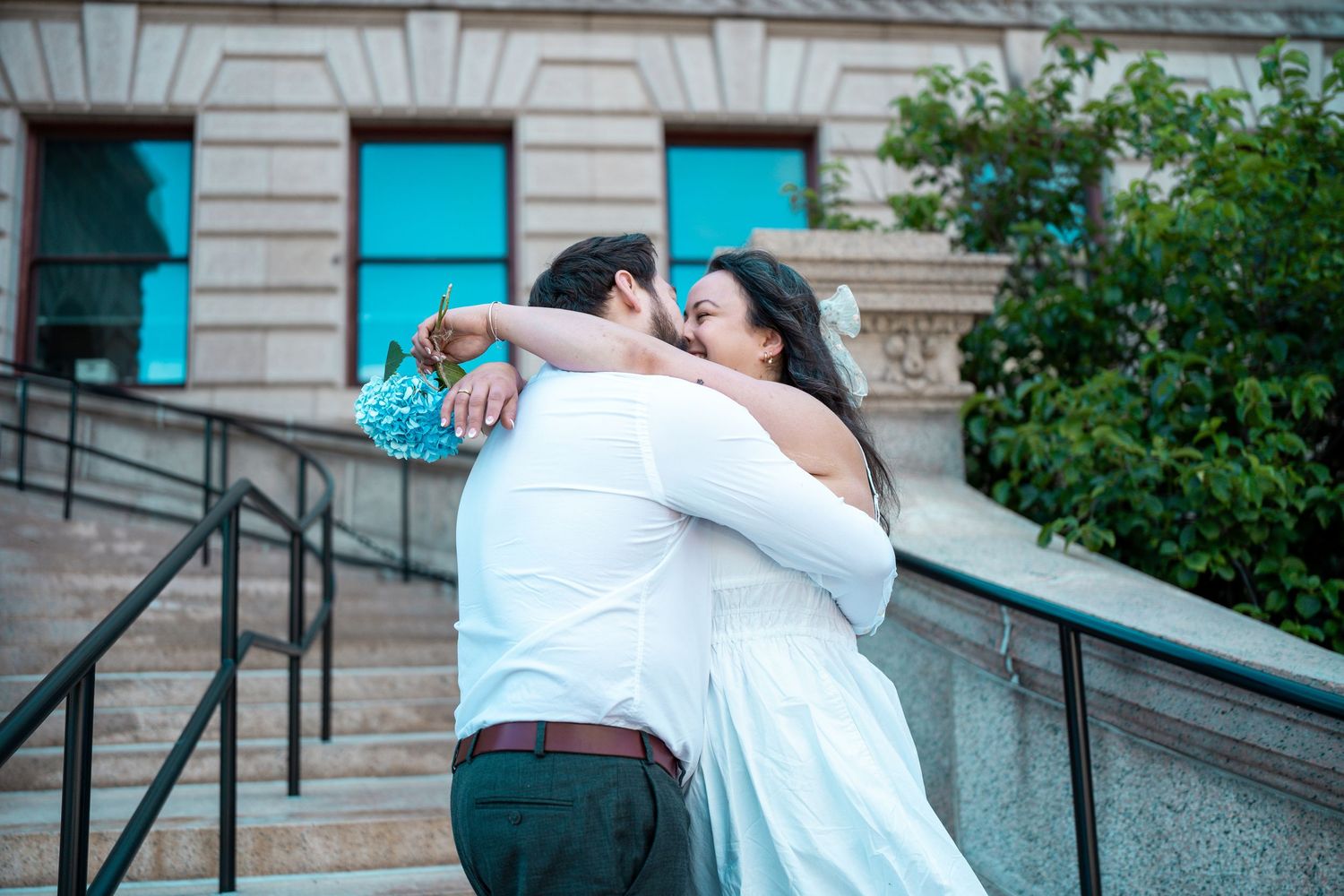 Bride and groom embracing on stone steps outside a building, holding a blue bouquet on their wedding day. Elopement at Worcester City Hall.