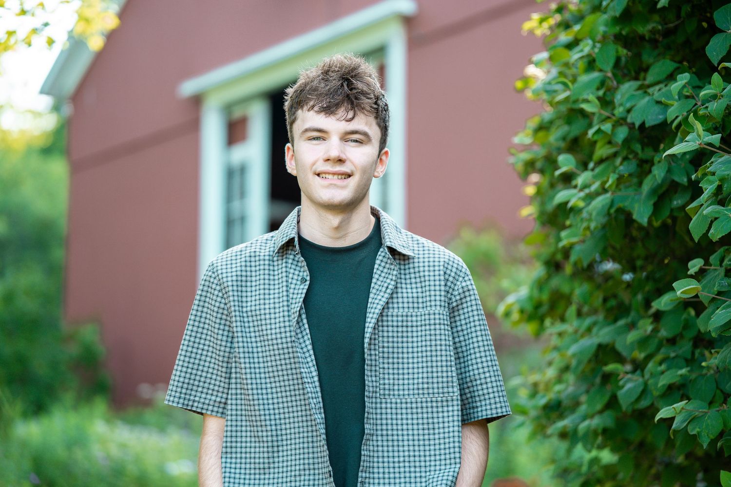 Smiling young man in plaid shirt standing outdoors near red building and green foliage.
